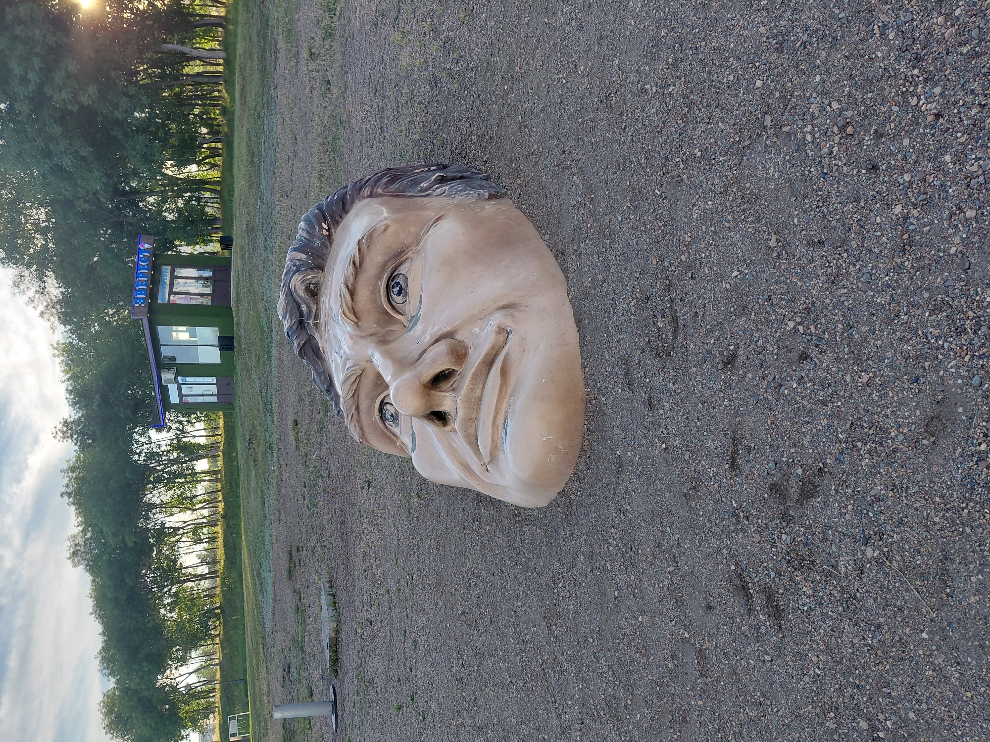 A large, realistic sculpture of a man's head, made of light brown material, is partially buried in gravel. The head is positioned in the foreground, facing slightly to the left of the camera. The sculpture is detailed, with wrinkles on the forehead and around the eyes, and appears to be weathered. In the background, a row of green trees runs horizontally across the image, with a small, modern-looking kiosk or building situated amongst them. The kiosk has a blue neon sign with Russian text that appears to read "Энергия" (Energy) or similar. The ground is covered in loose gravel and some sparse, dry grass. The sky above is partly cloudy with the sun beginning to set, casting a warm light on the scene. The overall impression is one of an outdoor sculpture in a park-like setting, possibly a public space. This is the sculpture "The Head" by Ivan Svetchnikov located in Magnitogorsk, Russia.