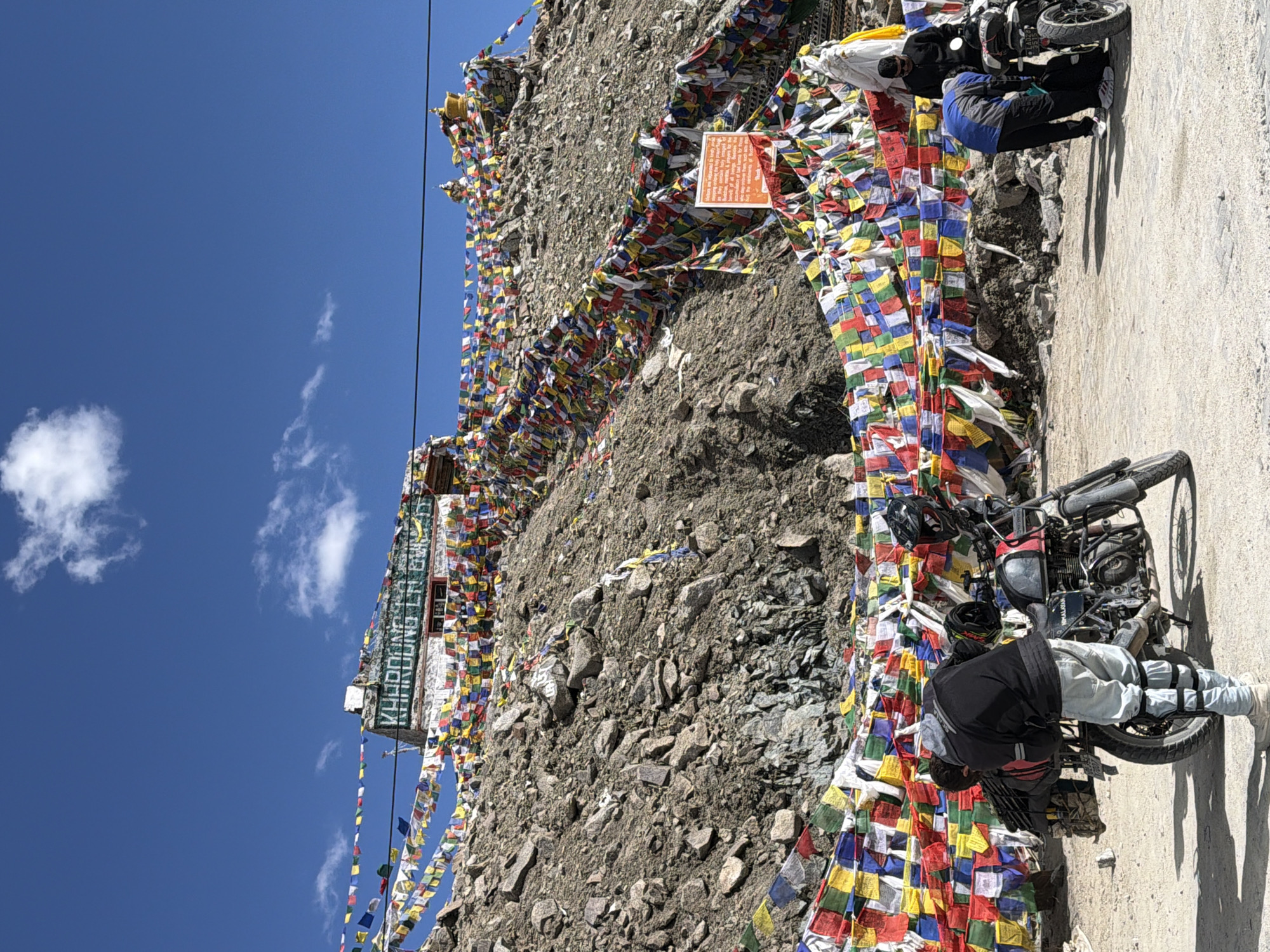 At Khardung La Pass, a high-altitude mountain pass in Leh, India, prayer flags in various colors are draped across a rocky slope leading up to a building. The building, likely a rest stop or administrative outpost, has a sign reading "Khardung La Pass". A group of people are gathered near motorcycles, some tending to them, others observing the surroundings. The ground is dusty and appears to be a road or parking area. The sky is clear and blue, with a few white clouds.