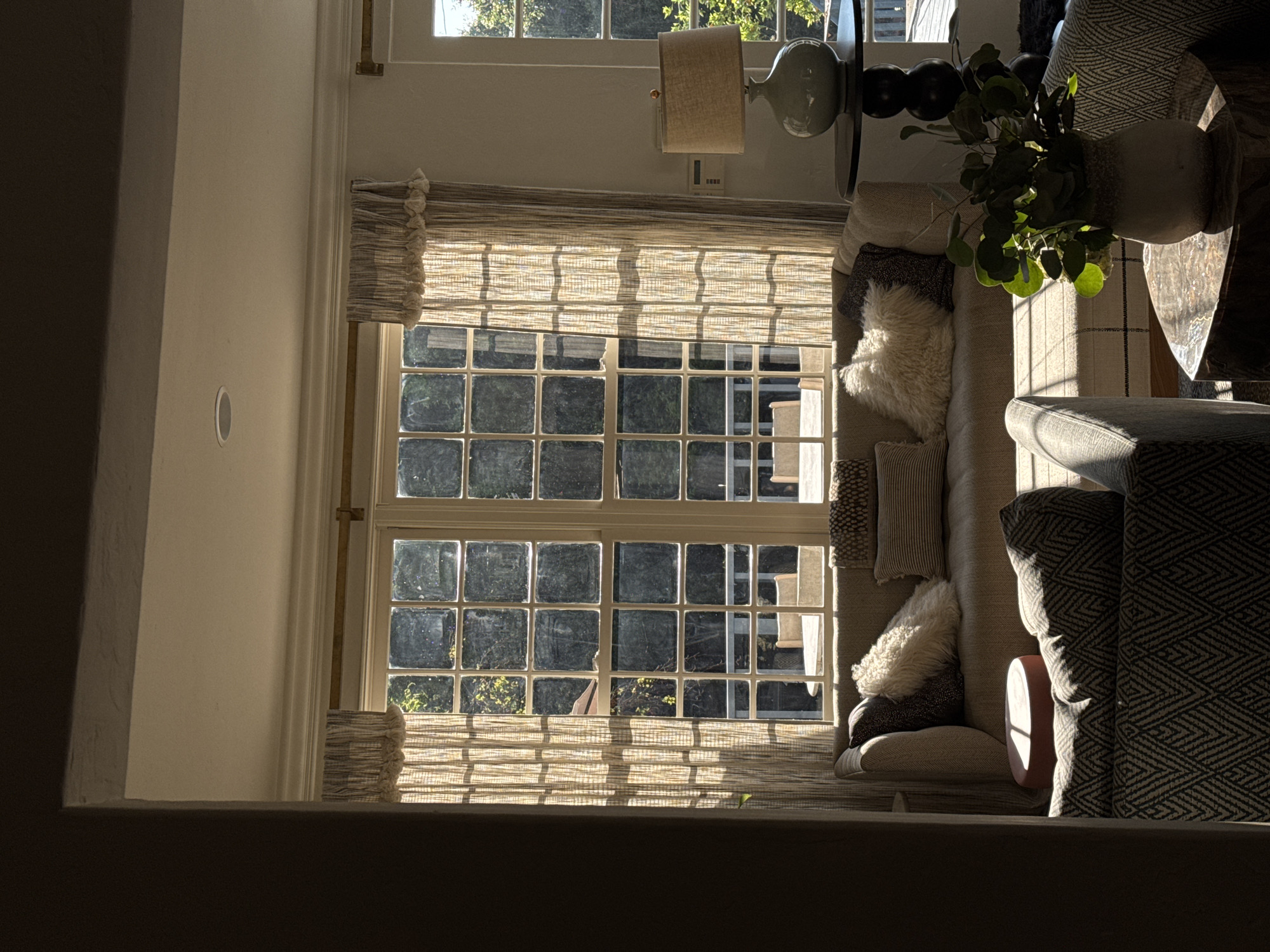The image displays an interior residential living space in Los Angeles, United States of America, illuminated by direct natural sunlight. No individuals are present.
The central feature is a large, multi-pane window unit with white frames, overlooking an outdoor patio or balcony containing furniture and surrounded by green foliage. To the right of this, a white-framed door or additional window unit is visible. Light-colored, vertically striped curtains with a ruffled top edge partially cover the windows.
Below the main window, a light-toned sofa or bench is positioned, adorned with multiple cushions, including dark patterned, light striped, and a prominent cream-colored, fluffy cushion. To the immediate right of the sofa, a dark, round, pedestal-style side table supports a light-colored ceramic lamp with a cylindrical fabric shade.
In the foreground, sections of two armchairs with a dark, geometric zig-zag pattern are visible. Between these armchairs, a dark, possibly stone, hexagonal side table holds a light-colored vase containing green leafy plants. A small, round, reddish-brown stool is partially visible in the left foreground, in front of the sofa.
Architectural details include white crown molding along the ceiling and a recessed light fixture. A dark doorway or archway frames the left side of the scene. A wall-mounted control panel is affixed to the wall beside the right window unit.