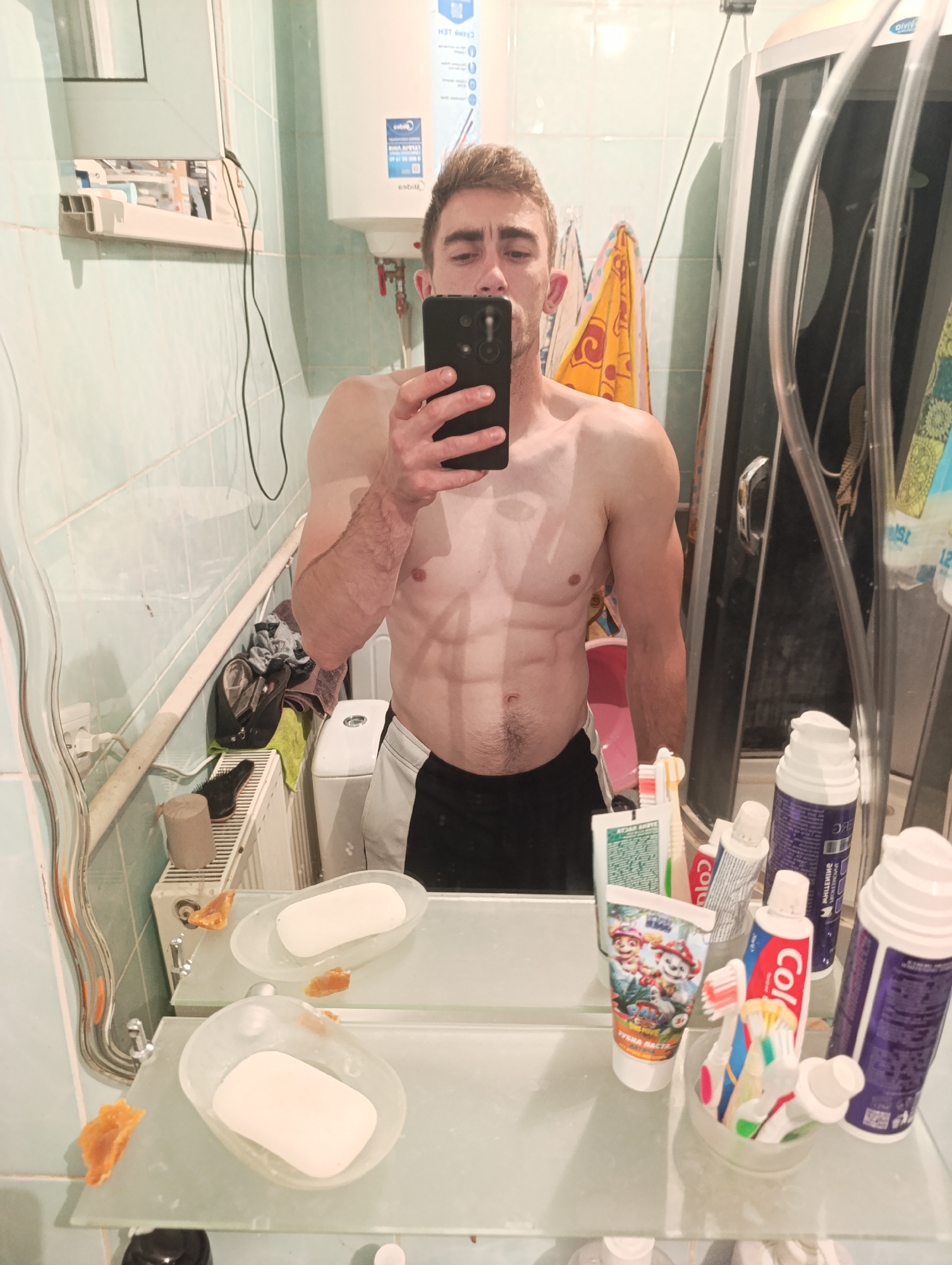 The image depicts a young man with visible abdominal muscles and defined arms posing shirtless in a bathroom, holding up a smartphone to take a mirror selfie. He is wearing dark shorts with a white stripe. The bathroom appears to be in a residential dwelling, with tiled walls, a shower enclosure, a water heater, and various toiletries on shelves. Specifically, there are soap dishes with bars of soap, tubes of toothpaste (one with a Paw Patrol theme), toothbrushes in a holder, and aerosol cans of shaving cream or similar products. A towel is draped on a rack in the background. The lighting suggests artificial indoor lighting. The image does not contain any explicit information that would allow for the determination of its location within Fastiv, Ukraine.