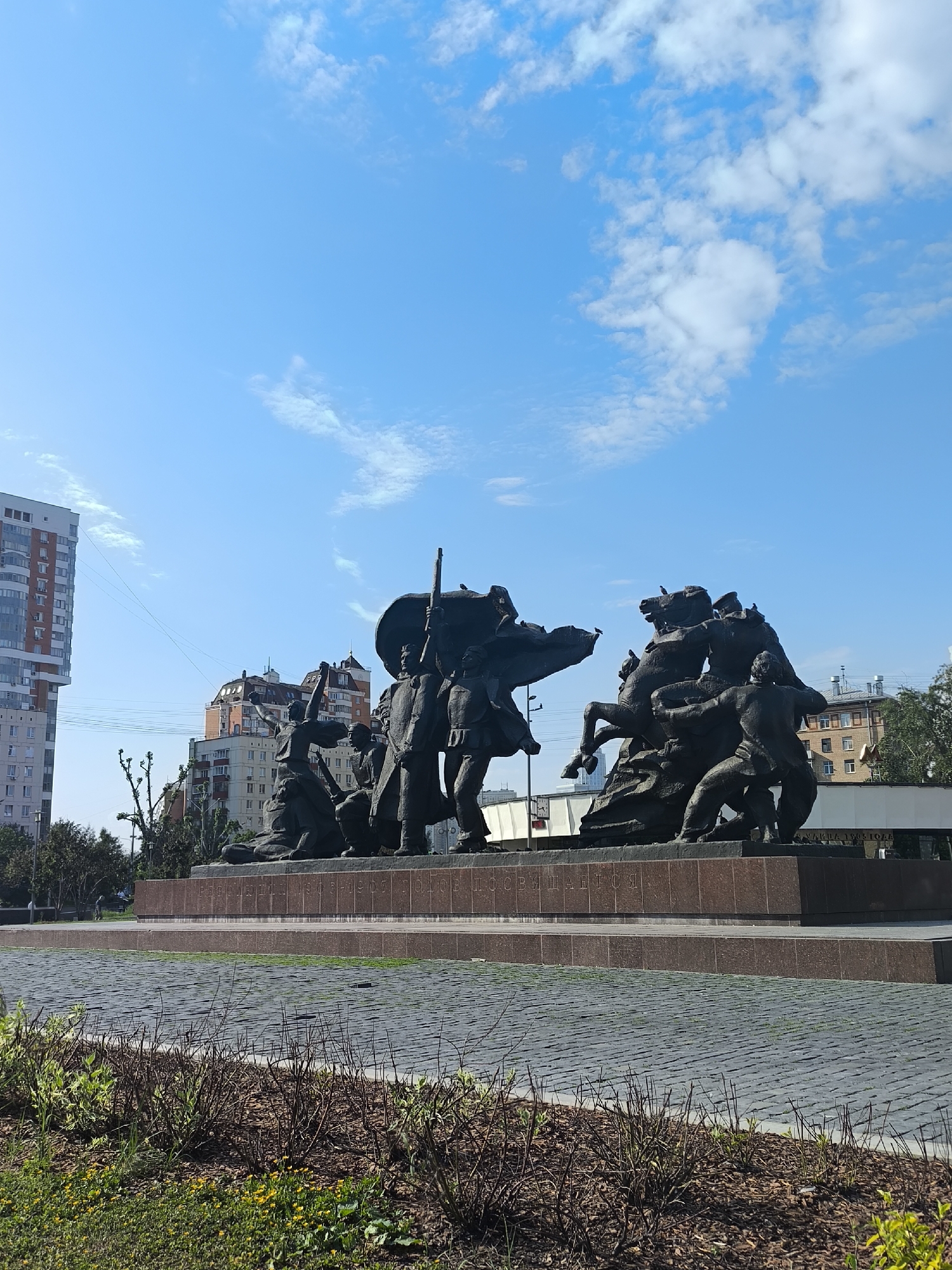 The image captures a bronze monument set against a backdrop of a clear blue sky dotted with sparse white clouds and urban buildings in Moscow, Russia. The monument, positioned on a tiered brick platform with a paved square surrounding it, features a group of figures engaged in what appears to be a struggle or battle. Among them, there are figures on horseback, and individuals holding what might be weapons or flags. The overall scene suggests a historical or commemorative theme, potentially related to a significant event or figures. The foreground includes some shrubbery, further grounding the monument in its urban setting.