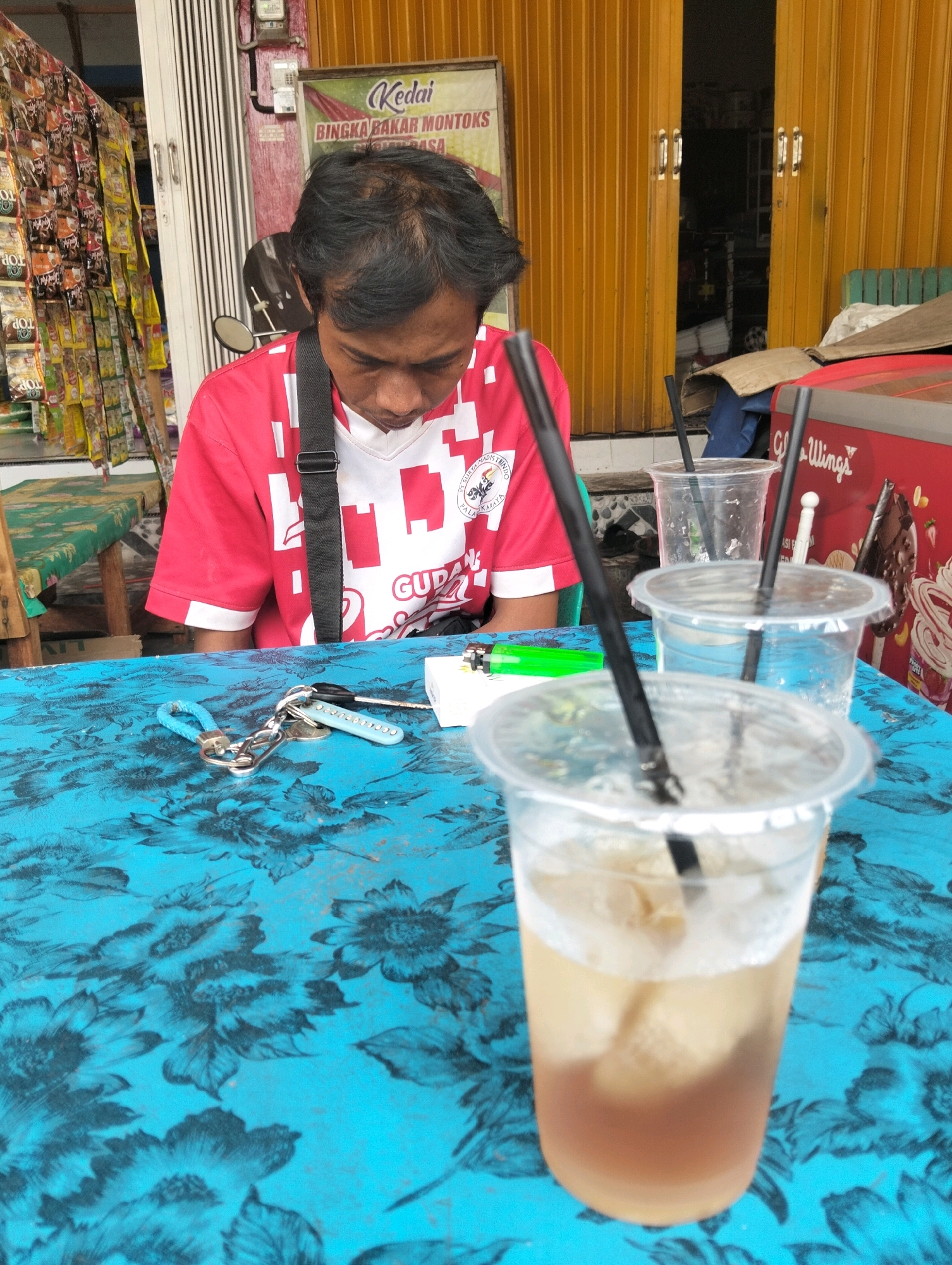 A man wearing a red and white shirt with a black strap across his chest is seated at a blue, floral-patterned table. He is looking down at a white object on the table, which appears to be a small box with a lighter next to it. Several plastic cups with black straws are also on the table, with one in the foreground filled with a light brown liquid and ice. Behind the man, a bright yellow corrugated metal door is visible, and to the left, racks of packaged goods are displayed. A sign above the racks reads "Kedai BINGKA BAKAR MONTOK." The scene suggests a casual setting at a roadside stall or small shop in Palangka Raya, Indonesia.