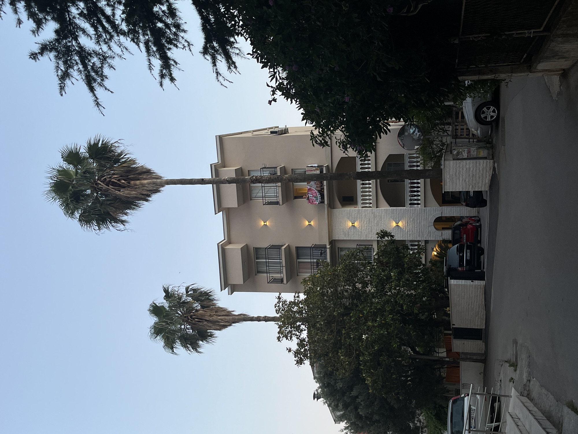 In Budva, Montenegro, an apartment building with a beige facade stands under a clear sky. Two tall palm trees rise in front of the structure, their fronds reaching towards the heavens. The building features multiple balconies with black railings, and warm-toned lights are mounted on its exterior. A small red car and two SUVs are parked in front of the building, and a silver car is visible on the left side of the image. Lush green trees and foliage surround the building, adding to the natural beauty of the scene.