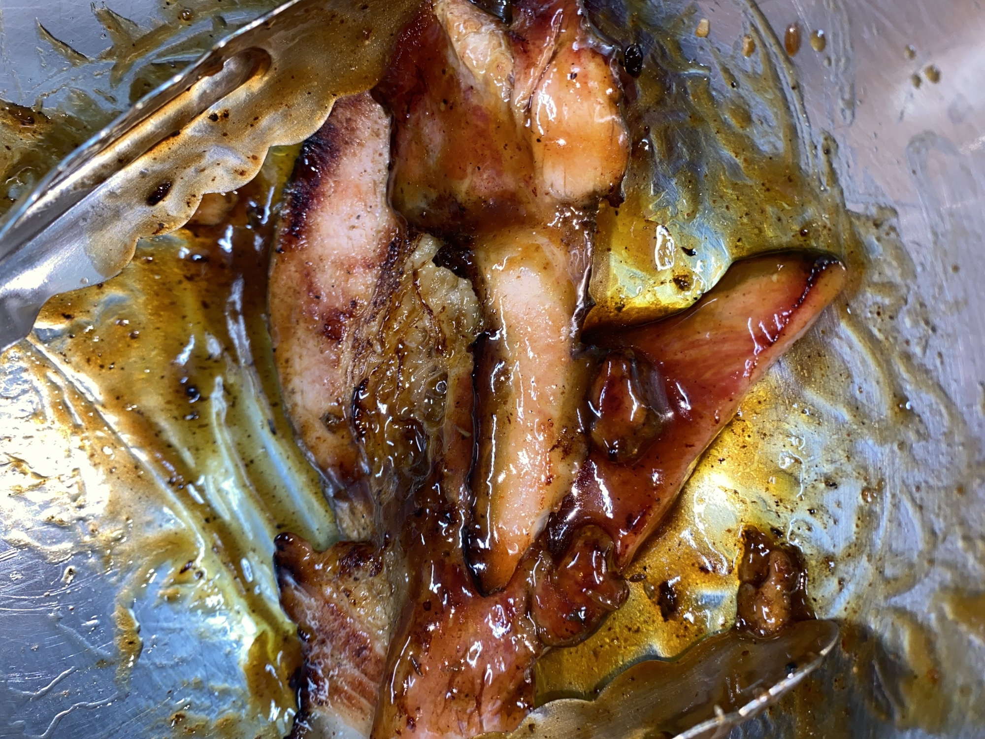 The image shows several pieces of cooked, glazed meat, possibly chicken or pork, held by metal tongs in what appears to be a reflective metal tray or container. The meat is coated in a dark, glistening sauce. No specific location or people are identifiable from the image.