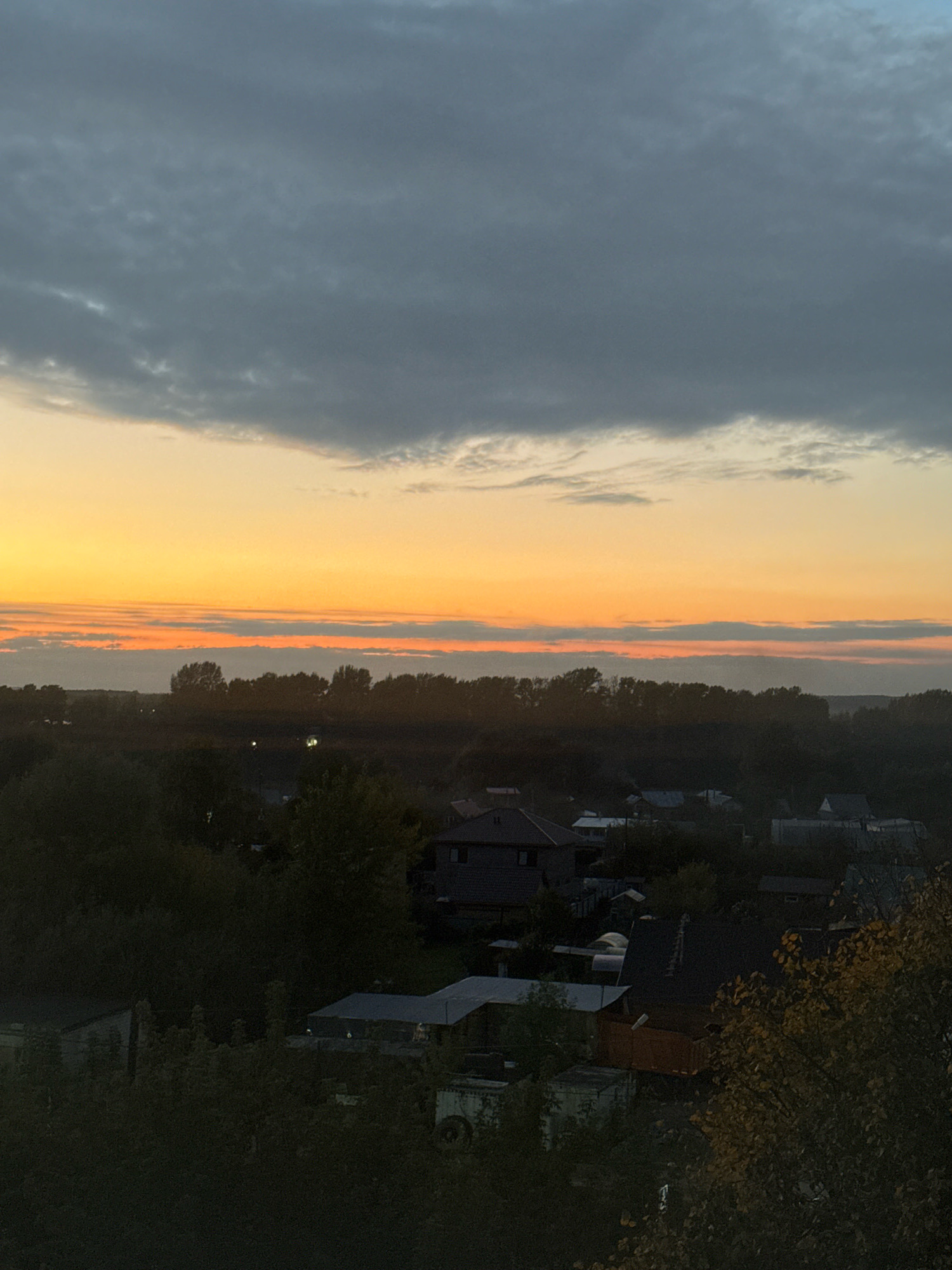 This is a vertical, high-angle, outdoor photograph capturing a sunset scene over a residential area in Chaikovsky, Russia. The sky is dominated by a gradient of colors, transitioning from dark gray clouds at the top to a vibrant orange and yellow sunset horizon, with thin, horizontal cloud layers illuminated by the setting sun. Below the sky is a dark silhouette of trees and the roofs of houses. The foreground features dense foliage and the upper portions of buildings, with some lights visible within the structures. The overall impression is of a quiet evening in a suburban or rural setting.
