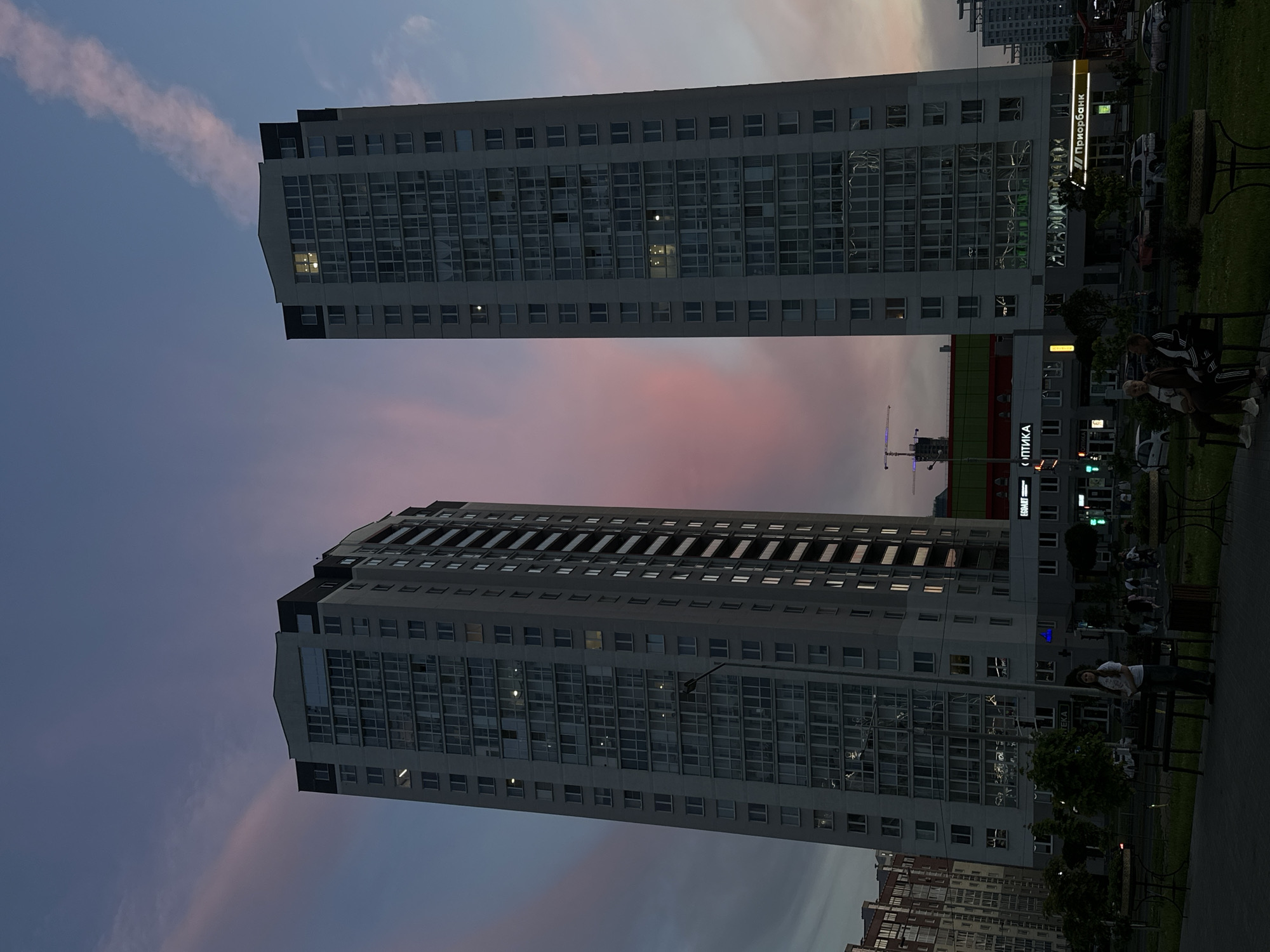 This media file presents an urban scene in Minsk, Belarus, likely captured during dusk or dawn, characterized by a sky transitioning from deep blue to soft pink and purple hues. Two modern, high-rise buildings, constructed from light-colored materials with numerous reflective glass windows, dominate the composition, appearing to stretch vertically across the frame.
Several people are visible on the right side of the image, near the base of one of the buildings and in an adjacent paved area. A woman in a white t-shirt stands by a dark bench, while two other individuals are seated on another bench, one prominently wearing a striped tracksuit. Other figures can be seen in the background, casually walking or standing, suggesting everyday activity in a public or residential space.
The scene features green spaces with grass and trees, bordering paved walkways and a street where cars are parked. A construction crane is faintly visible in the distance, hinting at urban development. Artificial lights from windows and commercial signs illuminate parts of the buildings.
Visible text provides strong location cues. On the right building, a prominent illuminated sign reads "Приорбанк" (Priorbank), a well-known Belarusian bank. Below it, parts of other signs are visible, including "ОПТИКА" (Optika, for optics or optician) and text that appears to be "OSMART". On the lower section of the left building, the partial text "ТЕКА" can be seen. The overall atmosphere appears calm and peaceful.