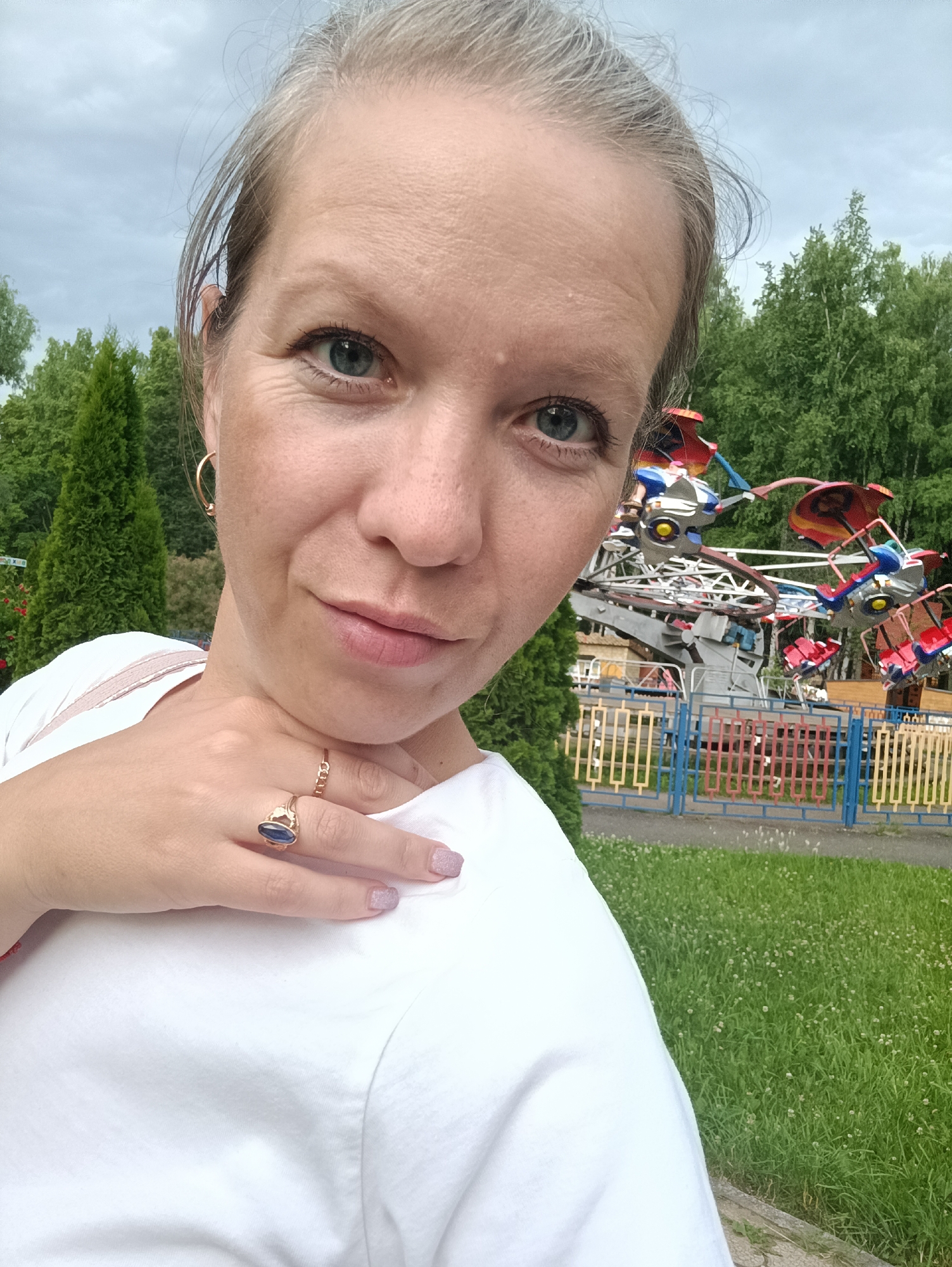 A woman is taking a selfie in an amusement park. She has light brown hair pulled back and is wearing a white t-shirt. She is wearing a gold hoop earring in her left ear and two gold rings on her right hand, one of which has a dark blue stone. Her left hand is resting on her neck. In the background, there is a green lawn with small white flowers, a colorful fence, and an amusement park ride with multiple colorful seats. Behind the ride, there are trees and a cloudy sky. The location is the amusement park in the city of Chaikovsky, Russia.