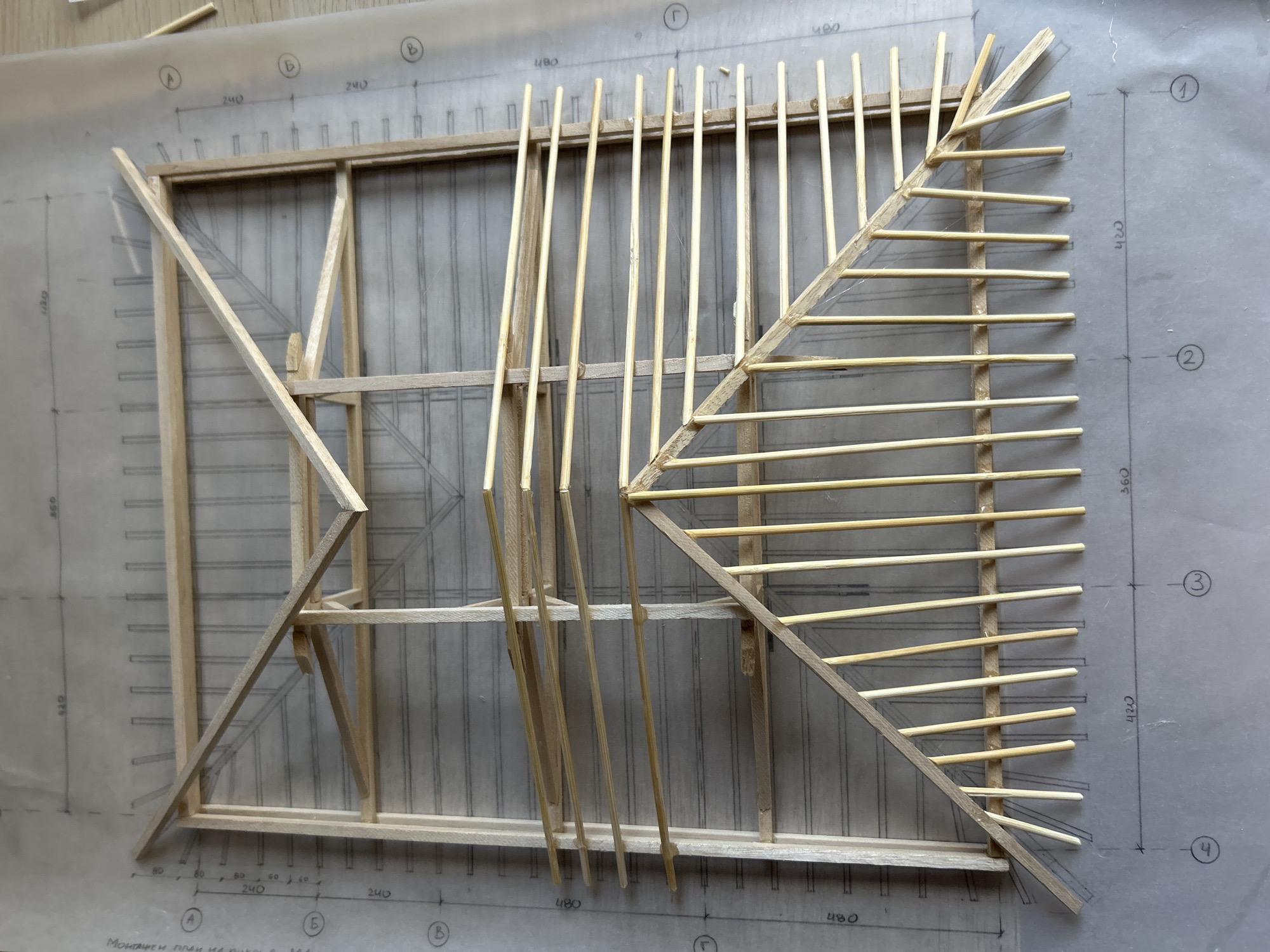 The media file displays an architectural model, meticulously crafted from slender, light-colored wooden sticks, resting on a translucent architectural drawing. The scene is likely an indoor setting, possibly a design studio or workshop, in Sofia, Bulgaria.
The model itself is a structural representation, primarily a rectangular frame. On the left side, it features an internal V-shaped bracing element. The right side is characterized by a long diagonal wooden member supporting a series of parallel, thin wooden rods that extend perpendicularly outwards, creating a fanned or rafter-like pattern. Another set of parallel rods spans the central part of the rectangular frame. Loose wooden sticks are visible in the upper left corner, suggesting either surplus materials or components for further assembly.
Beneath the model, the architectural drawing is visible through the translucent paper. It features a grid system with numerical dimensions such as "240", "480", "400", "360", "420", "80", and "60", likely indicating measurements in millimeters. The grid lines are labeled with Cyrillic letters "A", "Б", "В", "Г" and numbers "1", "2", "3", "4". A key piece of visible Cyrillic text reads "Монтажен план на роля", which translates to "Assembly plan of the role" or "Assembly plan for a part", explicitly indicating the purpose of the drawing. The image captures the model from a top-down, slightly oblique perspective, highlighting the intricate details of its construction and its alignment with the underlying plans.