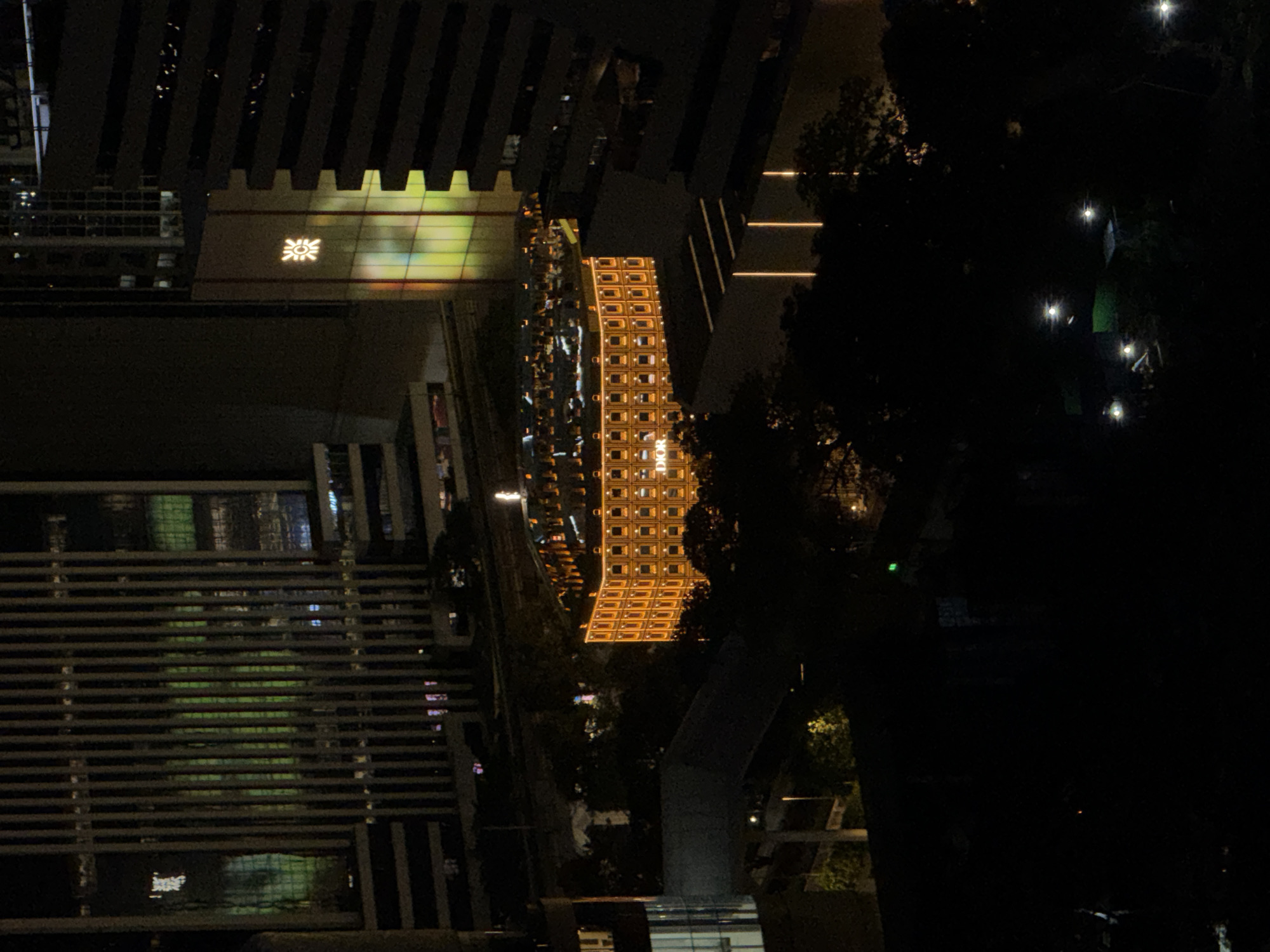 This is a nighttime aerial photograph of the Emporium, a luxury shopping mall located in the Phrom Phong district of Bangkok, Thailand. The mall is illuminated by golden lights and stands out against the darker urban landscape. Surrounding buildings are also visible, some with prominent architectural features and lit windows. The overall scene depicts a well-lit commercial hub within the bustling city.