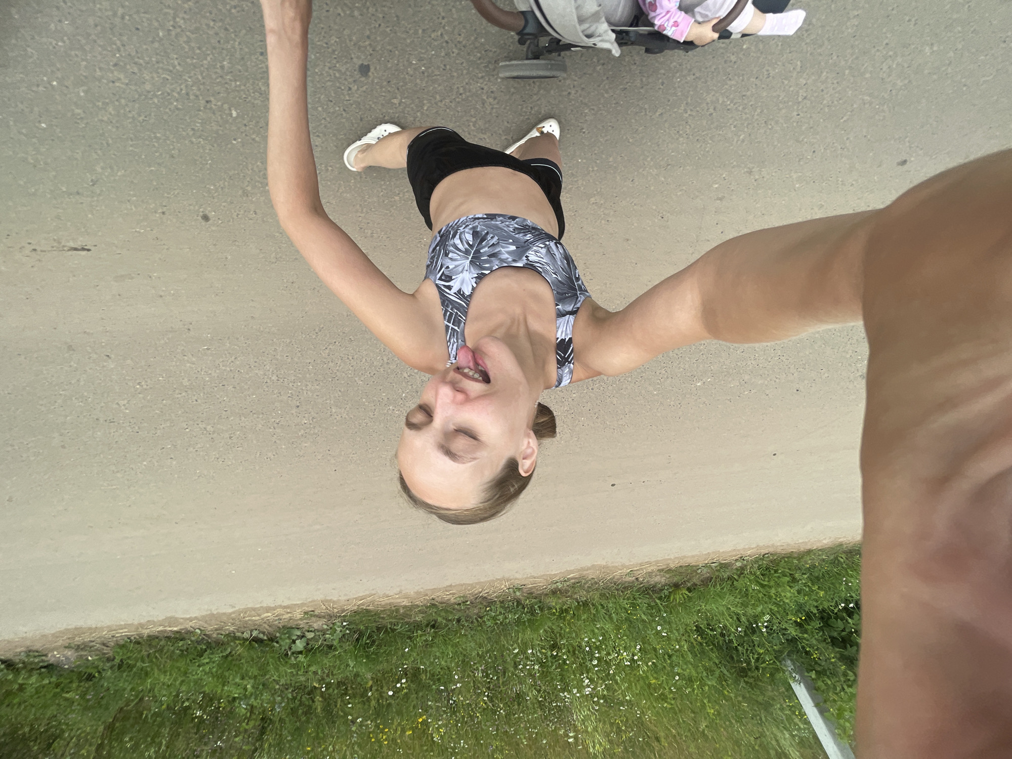A person is taking a selfie while standing on a dirt path next to a grassy area. The person is wearing a patterned sports bra and black shorts with white sneakers. They have their arms outstretched, and their eyes are closed with their tongue sticking out. In the background, there is a stroller with a child inside. The scene appears to be outdoors during the day, with green grass and a dirt path suggesting a rural or park-like setting. The weather appears to be fair. The location context provided is Podshivalovo, Russia.