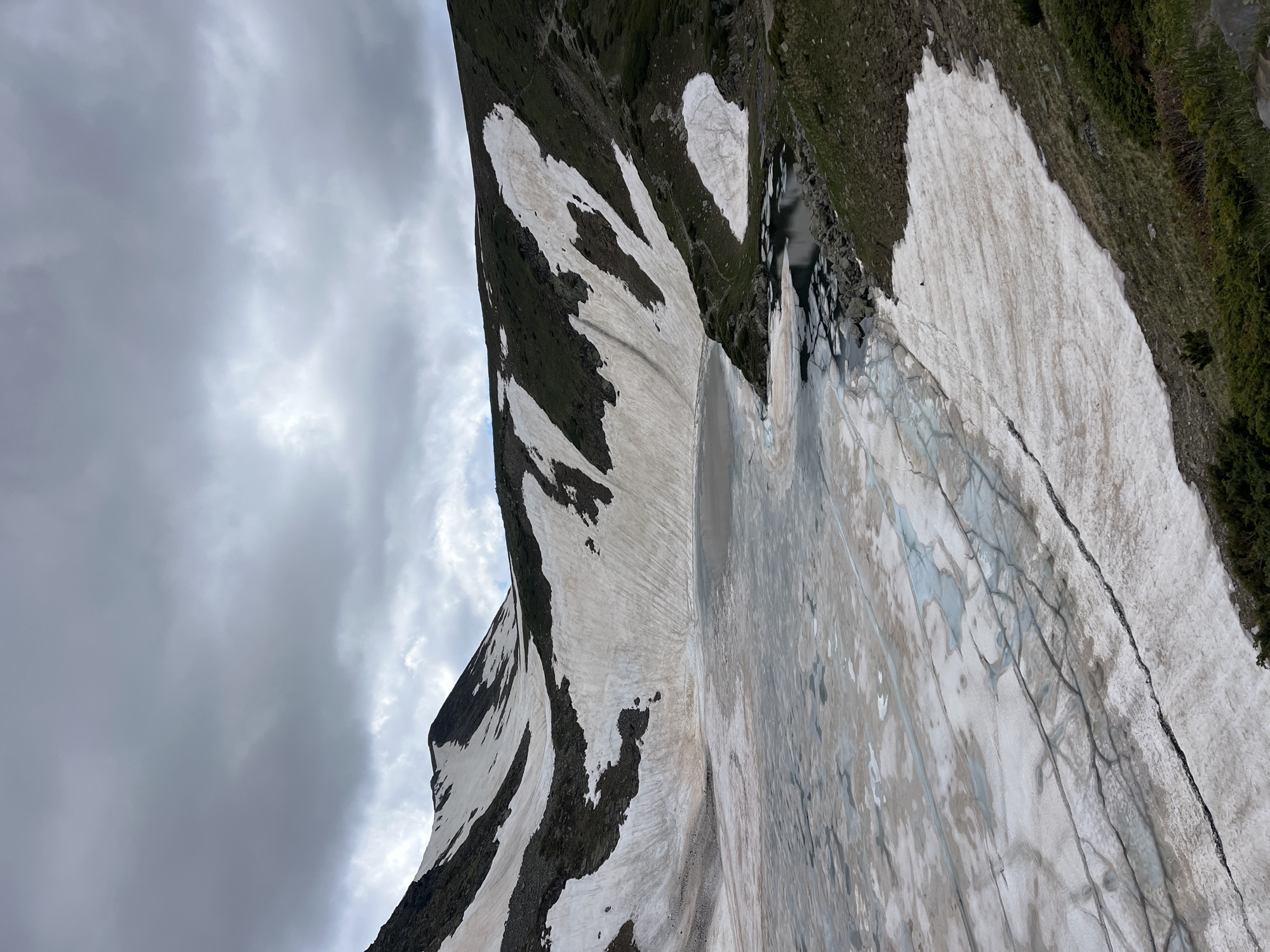 The image depicts a mountainous scene in Panichishte, Bulgaria, with a dramatic overcast sky dominating the left side. The terrain is rugged, with dark, rocky slopes interspersed with patches of snow. The snow is not uniformly white but shows streaks of dirt and meltwater, suggesting a thawing period. In the mid-ground, a body of water, possibly a lake or pond, is visible, with rocks scattered along its edge. The right side of the image shows a steep, grassy incline with sparse vegetation. The overall impression is one of a wild, untamed landscape under a cloudy sky. There are no people or man-made objects visible. The time of day is difficult to ascertain due to the overcast conditions, but it appears to be daylight. The weather is cool and potentially damp, with heavy clouds indicating a possibility of rain. The location is clearly mountainous, with the snow patches suggesting a higher altitude. No text is visible in the image.