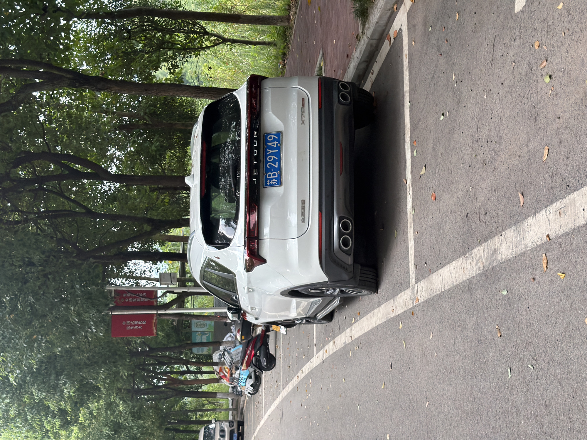 A white Jetour X70 Plus SUV is parked on the right side of a paved road, facing away from the camera. The car has a license plate that reads "苏B 29Y49". The rear of the vehicle features dual exhaust pipes. In the background, to the left, several motorcycles are parked. Behind the motorcycles, trees with dense green foliage are visible, as are two red banners attached to poles. The road is marked with white lines, and fallen leaves are scattered on the asphalt. To the right of the parked car, a red brick pathway runs parallel to the road, bordered by grass and more trees. The environment appears to be an outdoor setting, possibly a park or roadside area in Jiangjin, China.