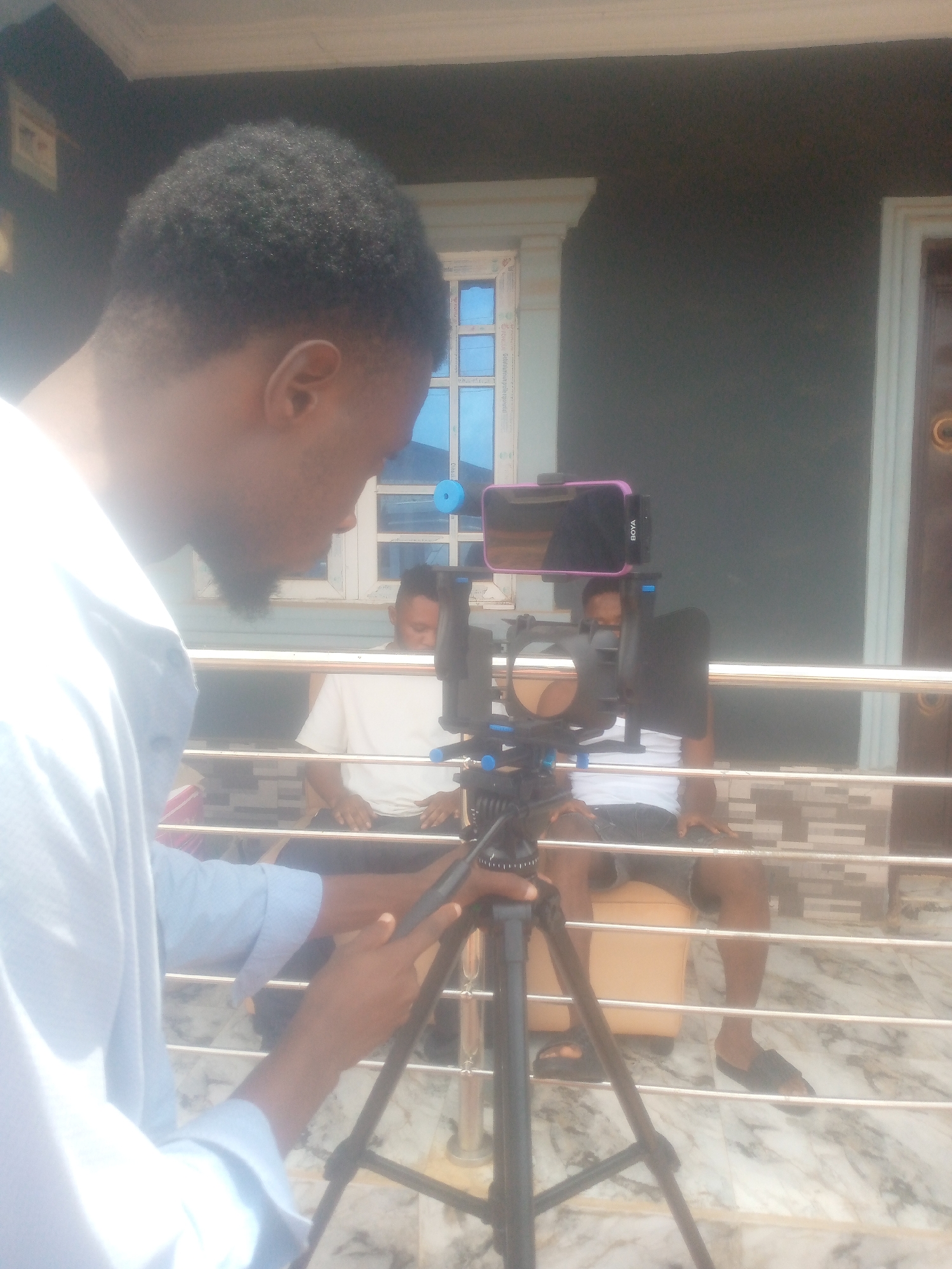 The image captures an outdoor scene in what appears to be a residential area in the city of Unknown, Unknown.
The foreground features a person holding and adjusting a black tripod. Atop the tripod sits a smartphone in a pink protective case. The smartphone seems to be equipped with a small microphone labeled "BOYA". Behind the tripod, there is a metal railing that seems to separate the foreground from the background.
Two individuals are seated in the background. Both are wearing white shirts and dark shorts. They are sitting on yellow stools.
The backdrop is a dark building with a window and a door. The window has a white frame and blue glass. The door is also white and features a small brass handle. The porch floor is a light gray marble.