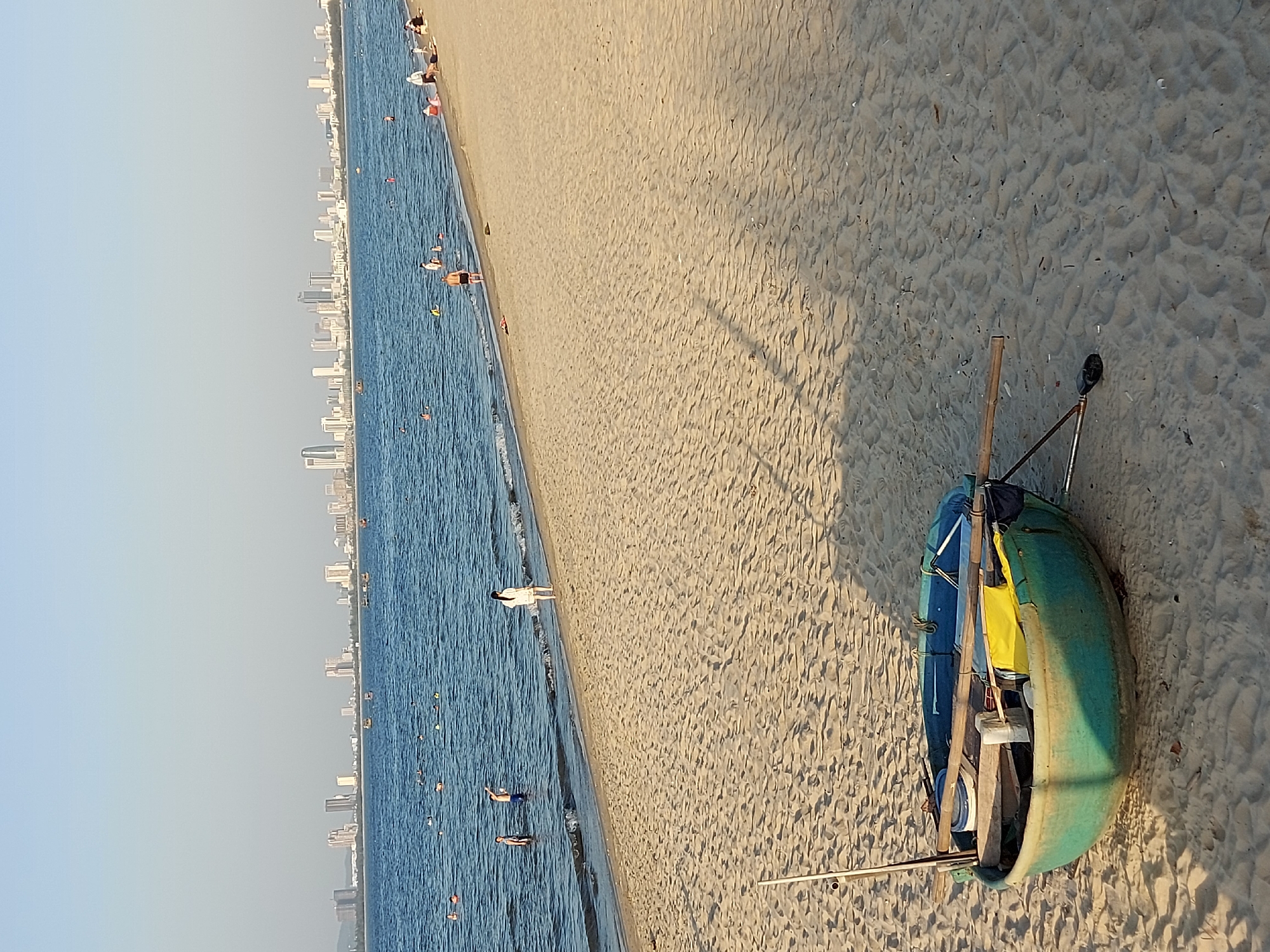The image depicts a coastal scene in Da Nang, Vietnam, with a sandy beach in the foreground and the city skyline in the distance. The ocean is calm, with several people visible swimming and wading near the shore. The beach itself is mostly empty, with the exception of a small, round boat resting on the sand. The boat is green with blue accents and appears to be made of fiberglass. It is equipped with oars and other fishing gear. The city skyline, visible across the water, is characterized by a cluster of modern high-rise buildings. The sky is clear and blue, suggesting a sunny day. The overall impression is one of a peaceful, seaside environment.