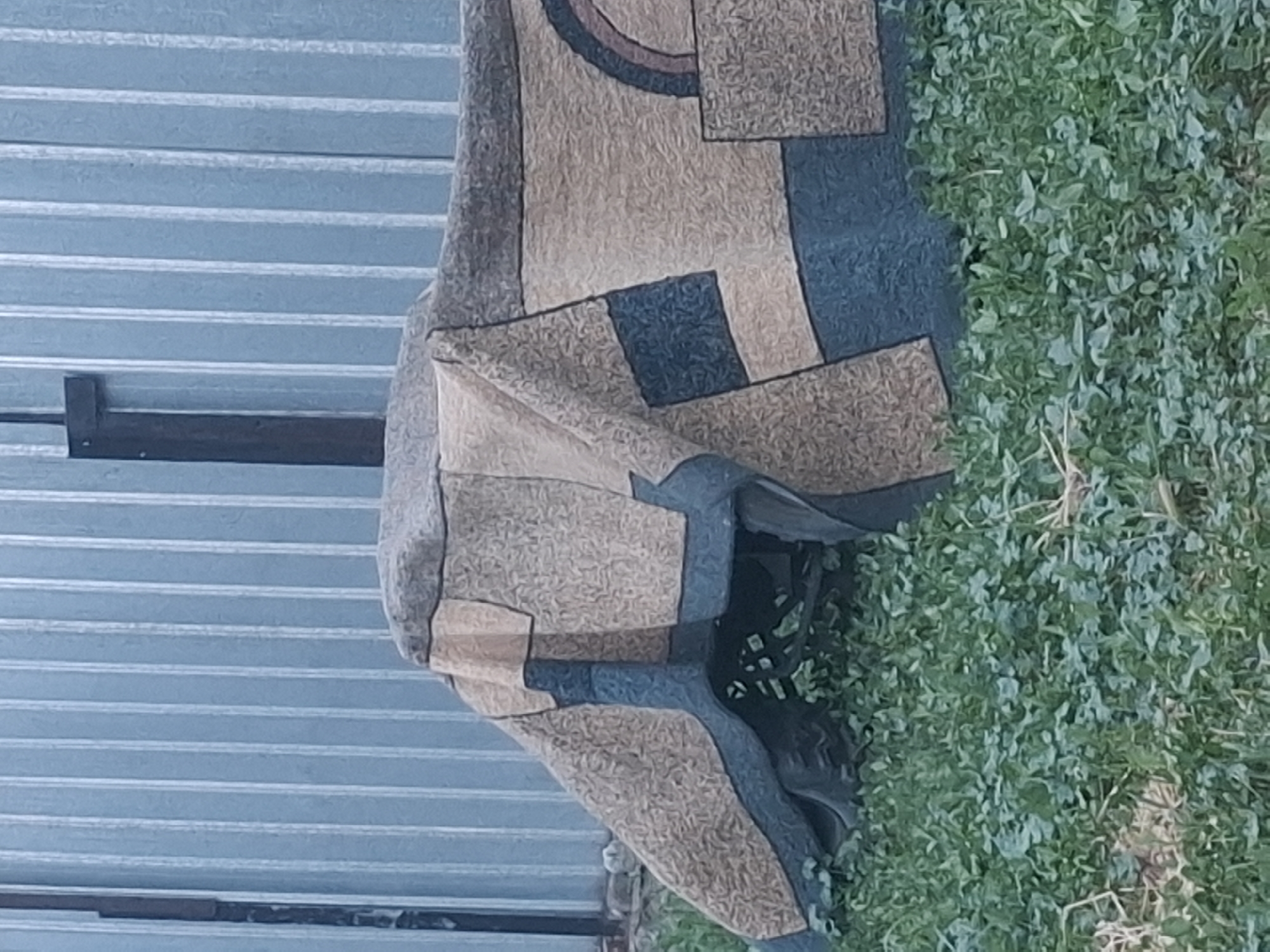 The image shows a close-up of a rolled-up rug with a geometric pattern. The rug is predominantly tan and dark blue, with some areas of brown and a curved line in a muted purple. The pattern consists of squares and rectangles of varying sizes, as well as a partial circle. The rug appears to be made of a textured material.
In the background, to the left, is a corrugated metal surface, likely part of a fence or wall, painted a light blue. A dark horizontal bar is visible attached to this surface. To the right of the rug, there is a patch of green grass with some dry blades of grass scattered throughout. The lighting suggests it could be daytime, possibly outdoors. The setting appears to be a casual outdoor area, possibly a yard or patio. There is no visible text in the image.