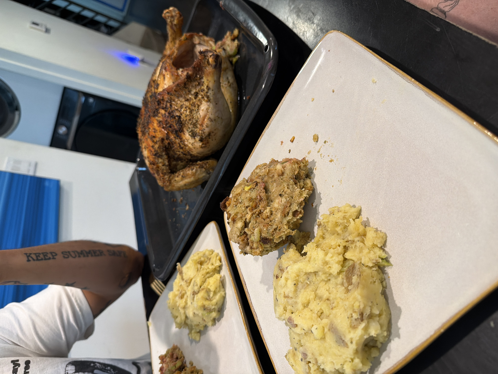 A high-angle shot depicts a cooked chicken in a roasting pan, accompanied by plates of mashed potatoes and stuffing. A person's arm with a tattoo reading "KEEP SUMMER SAFE" is visible in the lower-left corner of the image. The photograph appears to be taken indoors, possibly in a residential kitchen, based on the presence of appliances and furniture. Without further information, it is impossible to confirm the location within Los Angeles, United States of America.