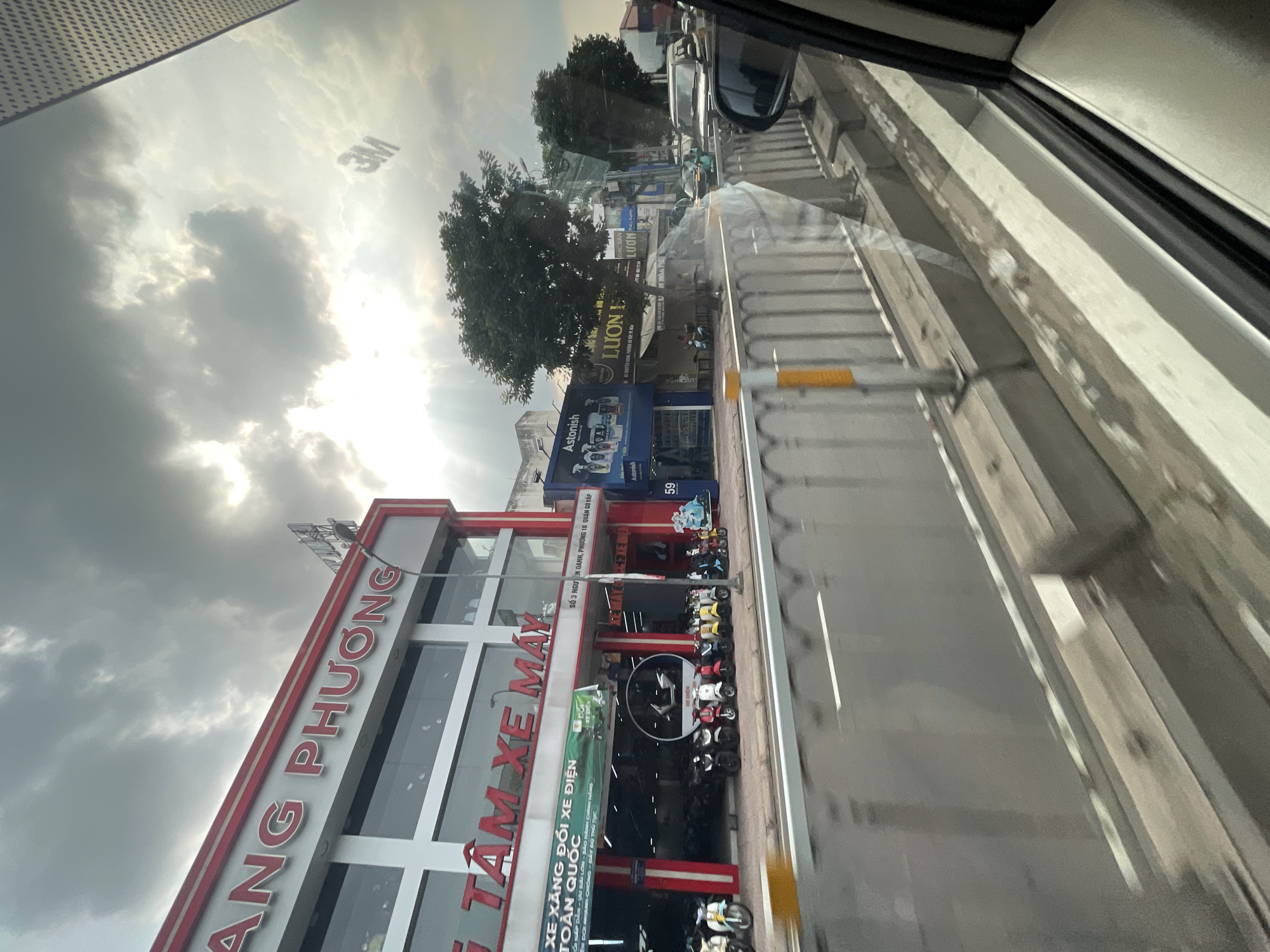 The image captures a street scene in Thuận An, Vietnam, under a cloudy sky with sunbeams breaking through. In the foreground, a busy road is visible, with a concrete barrier separating it from a sidewalk. On the sidewalk, a row of parked motorbikes is displayed outside a brightly lit showroom. The showroom, with red and white accents, has large windows showcasing more motorbikes inside. Above the showroom's entrance, a prominent sign reads "NG PHƯƠNG" in large, red, three-dimensional lettering. Below this, another sign states "TÂM XE MÁY". Further down the street, several other businesses with various storefronts and signage are present, including "Astonish" and "LUƠN L", along with trees lining the sidewalk. A few vehicles are on the road, indicating moderate traffic. The overall impression is of a commercial area on a partly overcast day.