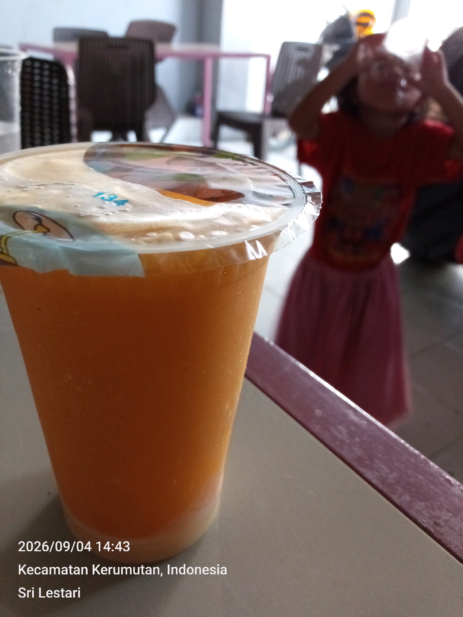 An orange beverage in a clear plastic cup with a sealed lid sits on a table. The lid is partially peeled back to reveal a light brown frothy layer. In the background, a blurred young child wearing a red shirt and a pink skirt is visible, with their hands raised near their face. The setting appears to be an indoor area with tables and chairs, possibly a cafe or food stall. The timestamp on the image reads "2026/09/04 14:43," and the location is identified as "Kecamatan Kerumutan, Indonesia, Sri Lestari."