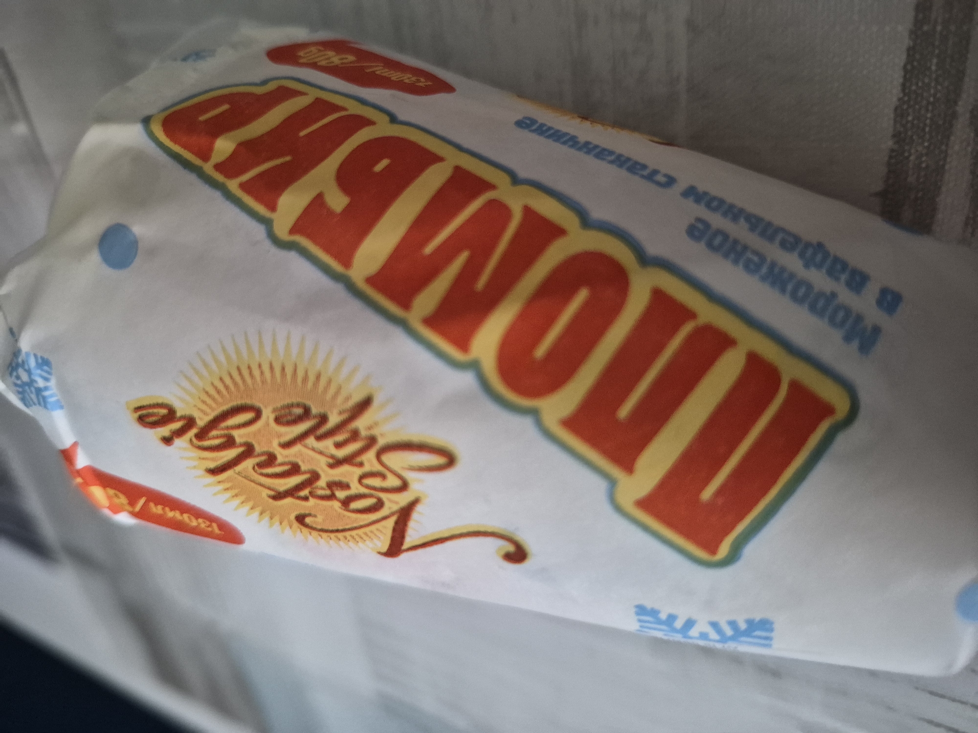 The image shows a close-up, angled view of an ice cream wrapper. The wrapper is predominantly white with colorful text and graphics. Dominating the center of the wrapper is large, bold, red and yellow text that appears to be in Russian. To the left of this text, there is a stylized sun graphic with the words "Nostalgie Style" in a script font. There are also some smaller blue circles and snowflake-like graphics on the wrapper. Towards the bottom right of the main text, there is smaller text which appears to also be in Russian, possibly indicating the product's description. The background is out of focus and appears to be a textured surface, possibly a tablecloth or a similar fabric. There is no indication of the location being in a city, nor are there any people or specific actions depicted. The overall impression is a product shot of packaged ice cream.
