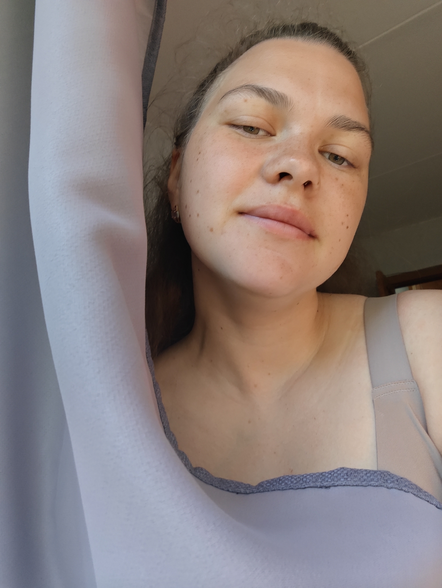 A woman with fair skin and freckles poses for a selfie. Her right arm is raised, with a light-colored curtain partially obscuring it. She is wearing a light gray tank top. The background is indistinct, but suggests an interior setting. It's impossible to determine the location in the city of Chaikovsky, Russia, from this image.