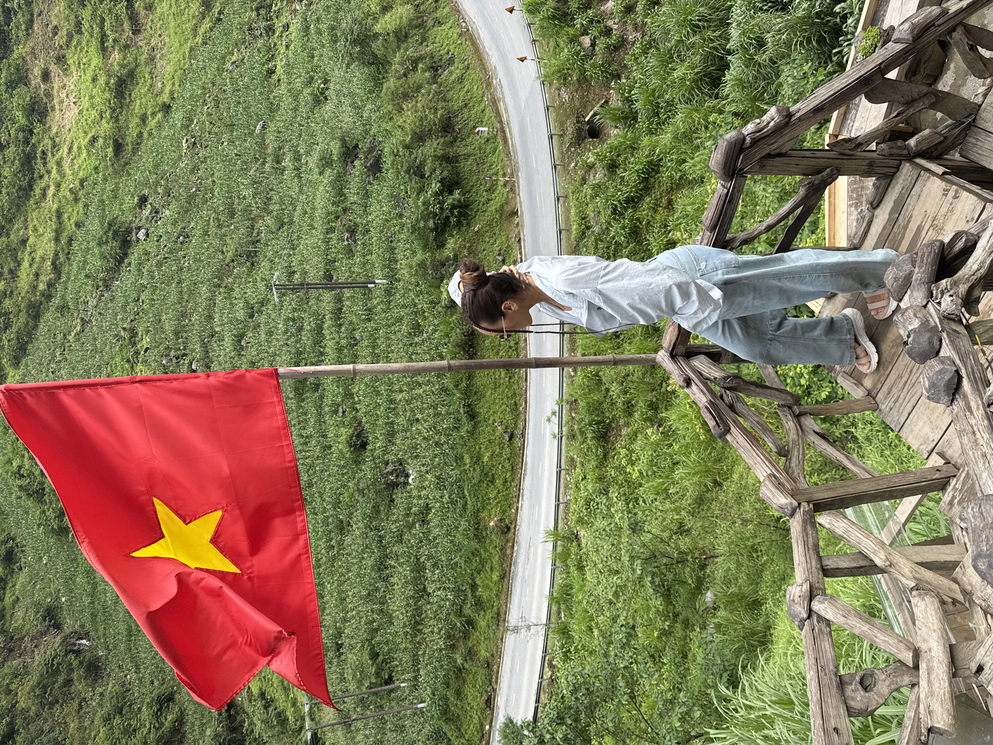 A woman is standing on a rustic wooden structure next to a road in Yên Minh Commune, Vietnam. She has her hair up, is wearing sunglasses, a light-colored shirt, and jeans. The woman is looking toward a large Vietnamese flag that is waving in the wind. The flag is red with a yellow star. The background consists of a steep, green hillside and a road winding through the landscape.