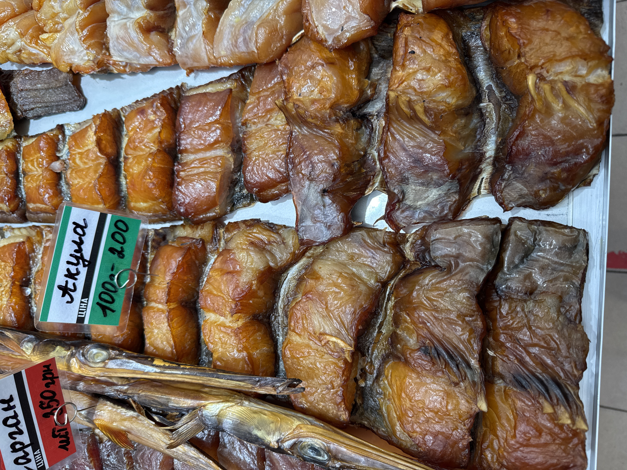 The image displays a close-up, overhead view of a market stall in Odesa, Ukraine, showcasing a variety of smoked fish. The fish are arranged in neat rows, with different types and cuts visible. Labels are attached to some of the fish, with Cyrillic script indicating product names and prices. One label reads "Сарван" and "150 грн," while another reads "Акула" and "100-200." The lighting suggests an indoor market setting, possibly a food hall or a dedicated fish market. The overall impression is one of abundance and variety in a local Odesa marketplace.