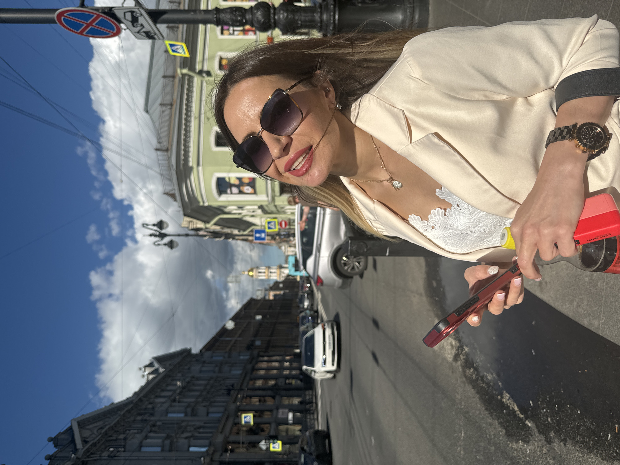The image shows a woman standing on a sidewalk in Saint Petersburg, Russia, holding a smartphone and a bottle of soda. She is wearing sunglasses, a white lace top, and a beige blazer. The background depicts a city street with cars, buildings, and a blue sky with clouds. A traffic sign indicating a tow-away zone is visible on a pole to the right of the woman. The overall scene appears to be a casual outdoor setting on a sunny day.
