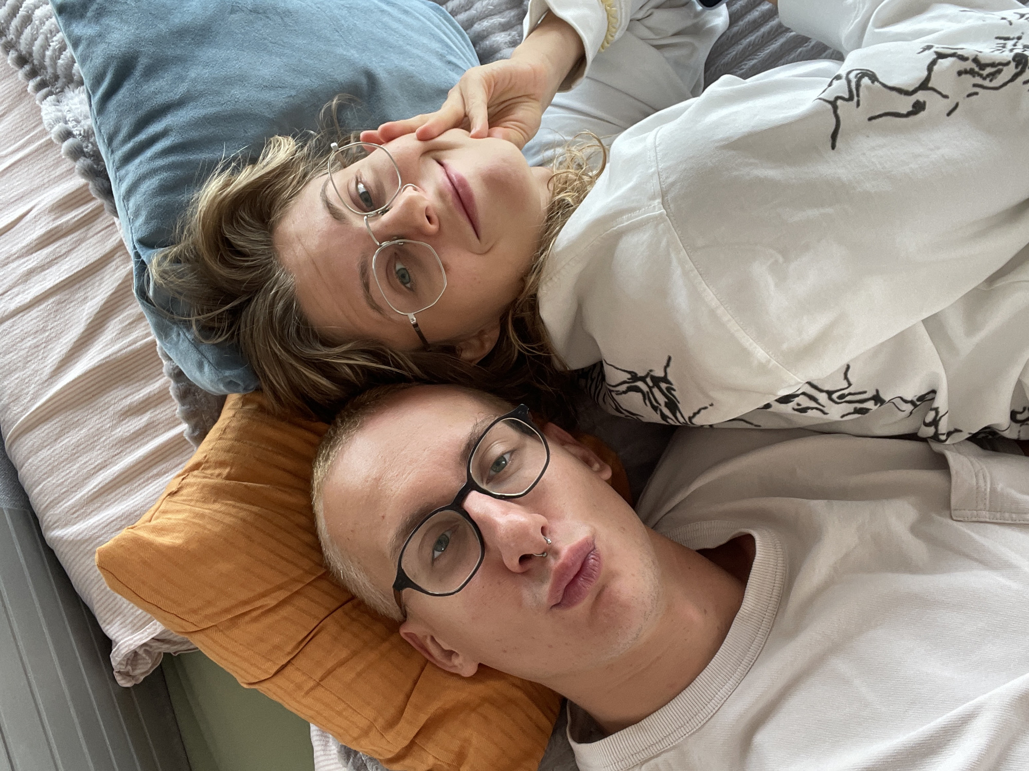 Two people are lying down, presumably on a bed given the presence of pillows and bedding. A person with short blond hair, wearing black-rimmed glasses and a silver nose ring, lies on an orange pillow. Next to them is a person with long, wavy blond hair, wearing round silver-rimmed glasses, lying on a blue pillow. The person with long hair is touching their face with one hand. The scene appears to be indoors. The location in Sestroretsk, Russia, is not discernable from the image.