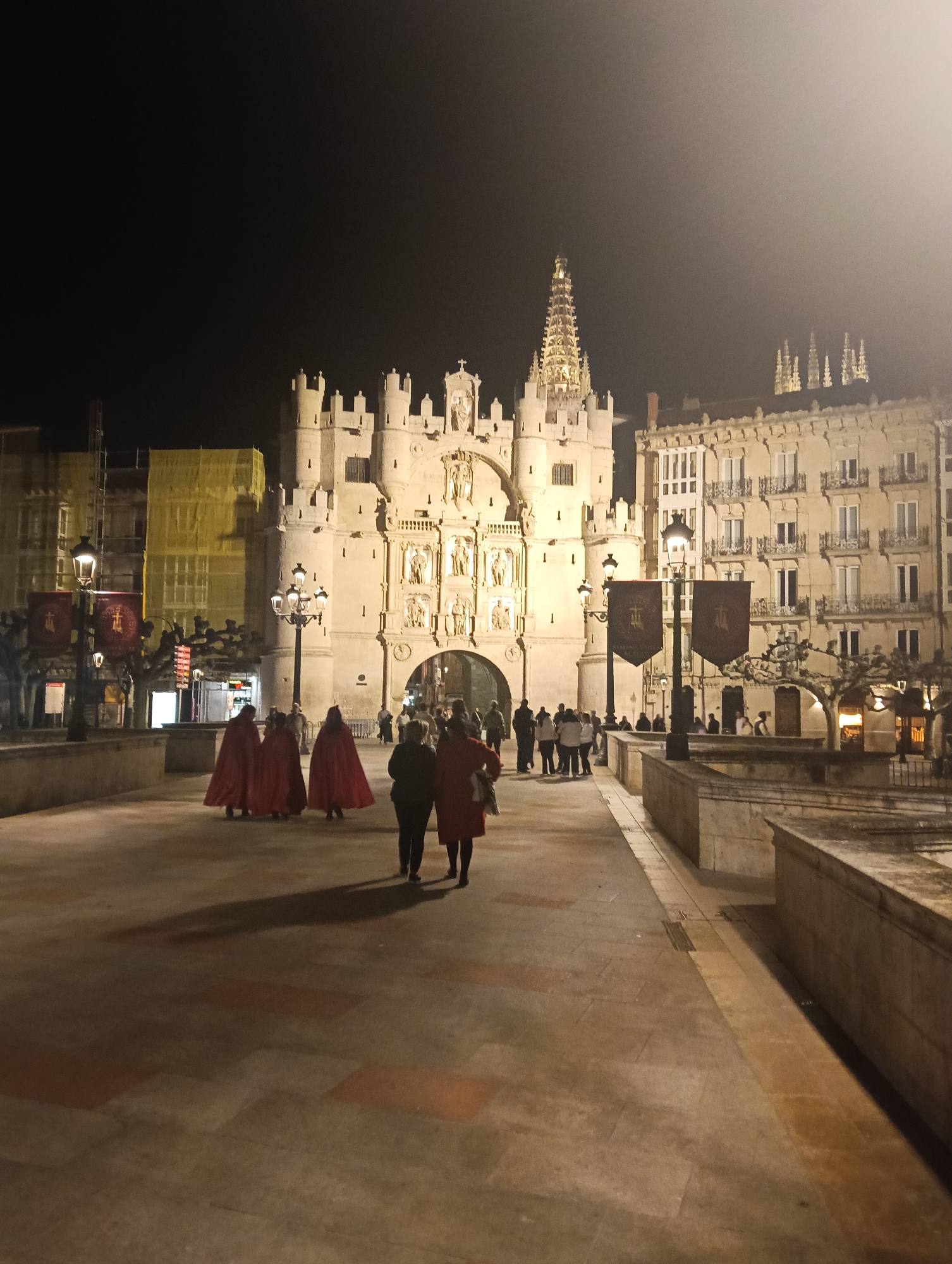The image captures a nighttime scene in Burgos, Spain, specifically the Santa Maria Arch. The arch, a prominent city gate, is brightly lit, showcasing its intricate architectural details and sculptures. To the left, a building is under renovation, covered with yellow scaffolding.
Several individuals populate the scene. A group of people in red cloaks are walking towards the arch on the left side of the frame. Two people are walking together on the right side of the frame. Other individuals can be seen milling about in the background, near the arch.
Banners are hanging from lampposts on both sides of the walkway, adding to the atmosphere. The ground is paved and appears damp or recently wet, reflecting the ambient light. Buildings and spires are visible in the background.