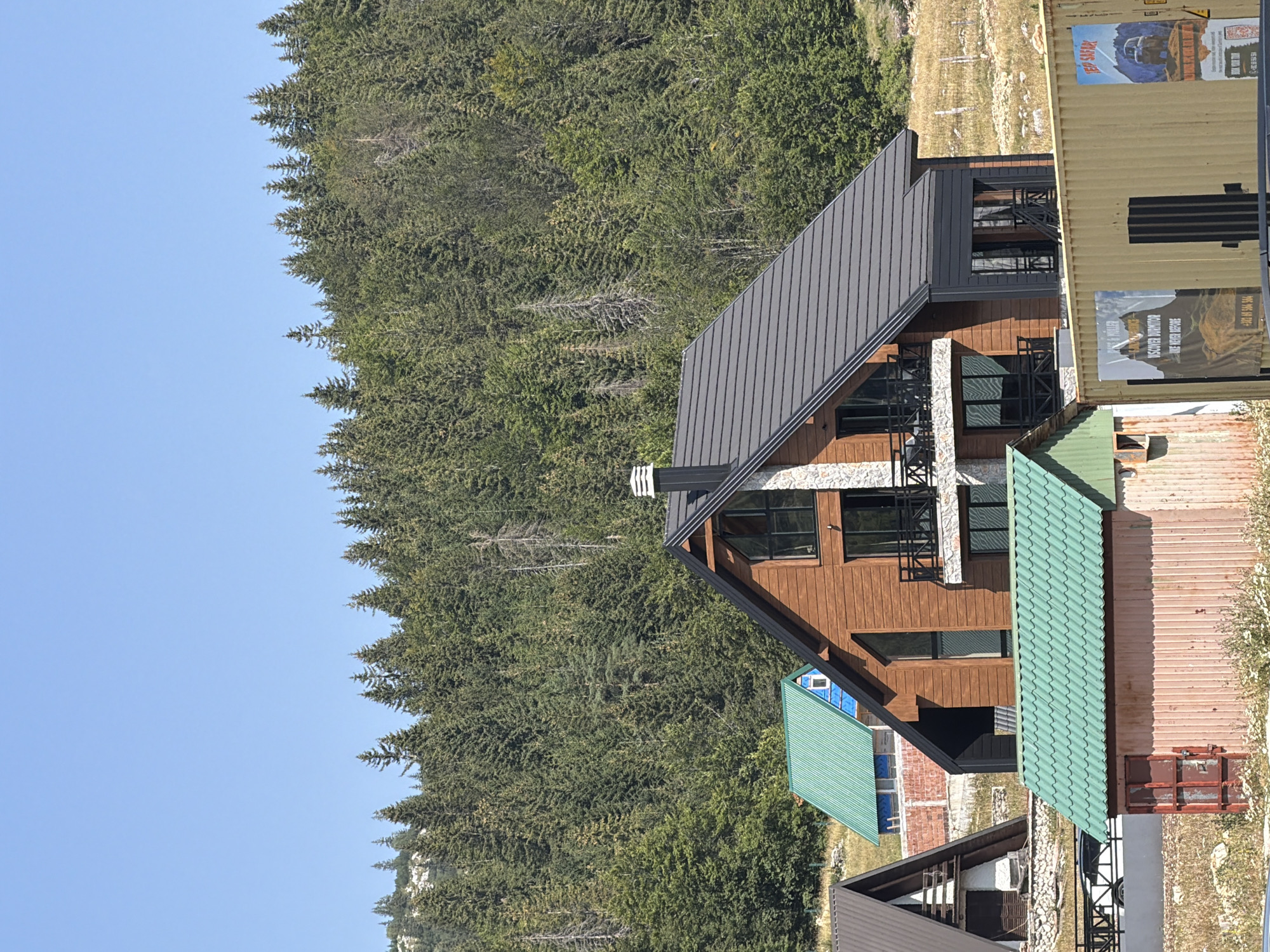 The image depicts a mountainous landscape with several buildings and a dense forest. In the foreground, there are three structures: a long, narrow building with a green corrugated metal roof and a rusty red door, and two houses partially visible to the left. One of these houses has a brown wooden facade and a dark gray roof. Further back, a smaller house with a bright green roof and blue trim is situated on a slope. The background is dominated by a vast, lush green forest covering the mountainside, with a clear blue sky above. The scene appears to be a rural or resort area, possibly in a mountainous region like Virak, Montenegro, given the architecture and natural environment. There are no people visible in the image.