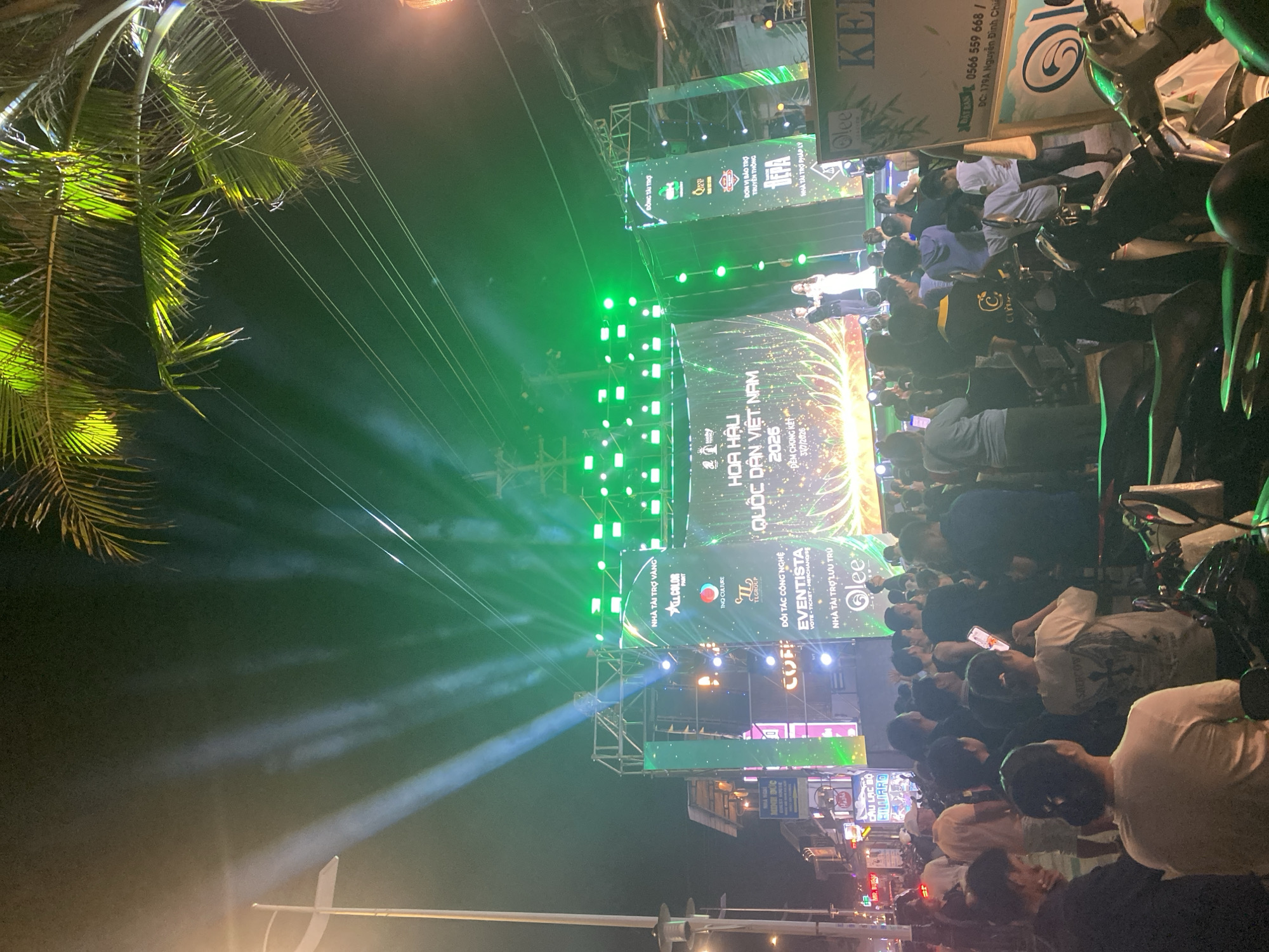 The image depicts an outdoor evening event, likely a beauty pageant, taking place in Mũi Né, Vietnam. A large stage with a prominent LED screen is the focal point, displaying the text "HOA HẬU QUỐC DÂN VIỆT NAM 2026" (Miss Nation Vietnam 2026). The stage is illuminated with green and white spotlights, and a palm tree is visible in the upper right corner, suggesting a tropical location.
On the stage, two individuals, a man in a dark suit and a woman in a white gown, appear to be participants or hosts, standing in front of the screen. The area in front of the stage is filled with a crowd of people, many of whom are holding up their mobile phones, possibly to record or take pictures. The crowd appears to be standing and gathered closely together.
To the right of the stage, a large sign with Vietnamese text and a logo is visible, with part of the text reading "DC: 179A Nguyễn Đình Chiểu". This indicates the location, Nguyễn Đình Chiểu street, a prominent street in Mũi Né, known for its tourism and businesses. Several motorcycles are parked in the foreground, suggesting this event is taking place in a public area where vehicles are commonly parked. The overall atmosphere appears festive and energetic, with bright lighting and a large audience.