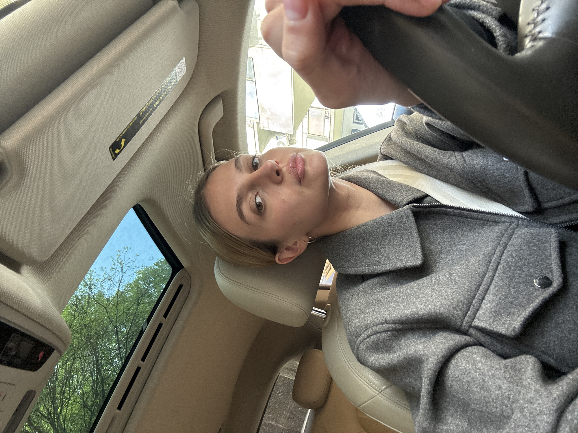 A woman is sitting in the driver's seat of a car. She is wearing a gray jacket and a seatbelt. Her left hand is on the steering wheel. The car has a sunroof. Outside the sunroof, trees and buildings can be seen, suggesting the car is likely in an urban area. The location is Tbilisi, Georgia.