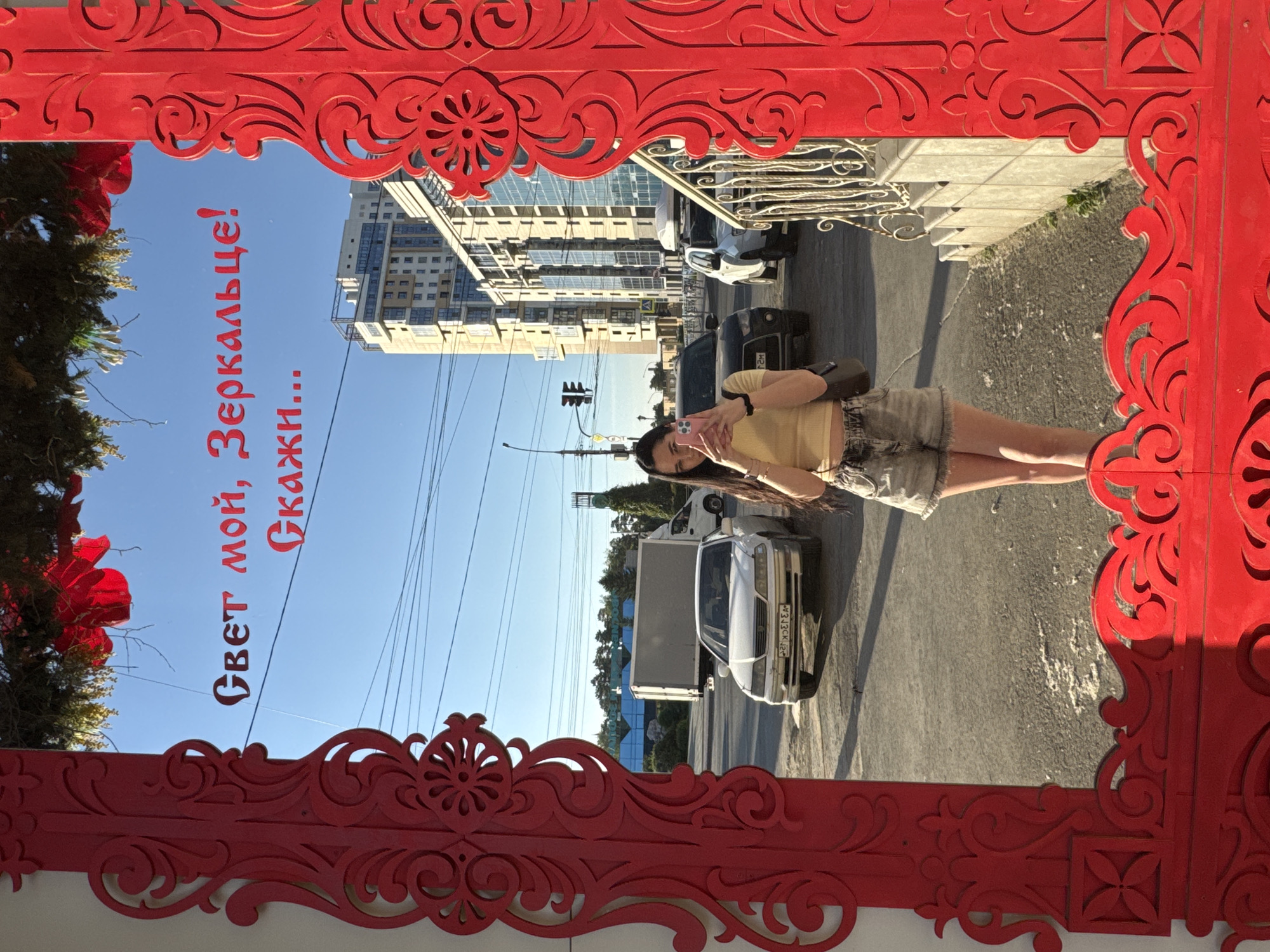 In a full-length mirror framed by ornate red decorations, a young woman is captured taking a selfie. She stands on a paved surface, possibly a sidewalk or street, with a clear blue sky above. The mirror reflects a city scene, featuring a modern, multi-story building with glass facades and balconies. Traffic lights, cars, and a large gray truck are visible on the street behind her, indicating an urban environment. The woman is wearing a yellow crop top and denim shorts. The red frame of the mirror includes carved floral and swirling patterns. The text "Свет мой, Зеркальце! Скажи..." which translates to "Light, my little mirror! Tell me..." is written on the mirror in red lettering. The presence of modern architecture and Russian text suggests this location is in Russia, likely in a city like Novosibirsk given the context of the building's design and style of lettering.