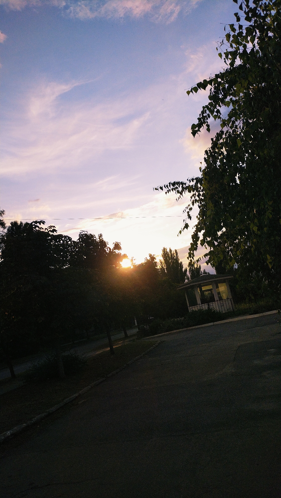 The image depicts an outdoor scene, likely a park or a street, in Horishni Plavni, Ukraine, during sunset. The sky is filled with soft pink and purple hues, with wispy clouds catching the light of the setting sun, which is partially obscured by dark silhouettes of trees. In the foreground, a paved asphalt surface suggests a road or walkway, with a curb bordering a grassy area. To the right, a small, white, gazebo-like structure with windows is visible, lit from within. The overall atmosphere is tranquil and serene, characteristic of a late evening in a residential or public area.