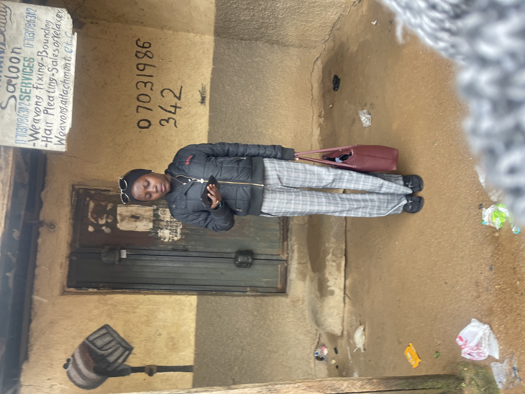 A young Black woman stands in front of a building with a stucco facade, the lower portion of which is a darker, rougher texture. She is wearing a black puffer jacket, plaid pants, and black sandals, with sunglasses perched on her head. She is holding a smartphone in her right hand and a maroon tote bag in her left. A sign with text about "Saloon Services" and phone numbers is visible on the wall above and to the right of her. Below the sign, a phone number is painted directly onto the wall. The ground is dirt and appears wet, with litter scattered around. The building has a metal door. A mural of what appears to be a lamp is painted on the wall to the left of the door. The image appears to have been taken in Rafin Par, Nigeria.