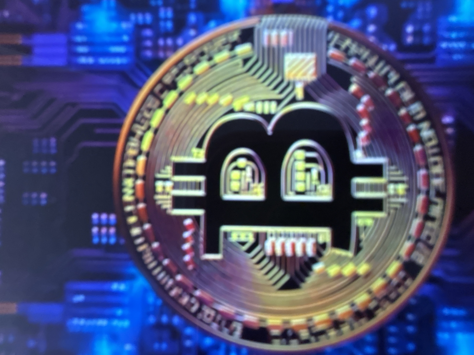 The image shows a close-up, slightly out-of-focus view of a Bitcoin cryptocurrency coin. The coin is gold-colored with a black "B" symbol in the center, characteristic of Bitcoin. The background is a blurred, dark blue digital circuit board pattern. There is no discernible location in the city of Gelendzhik, Russia, nor are there any people or specific actions depicted.