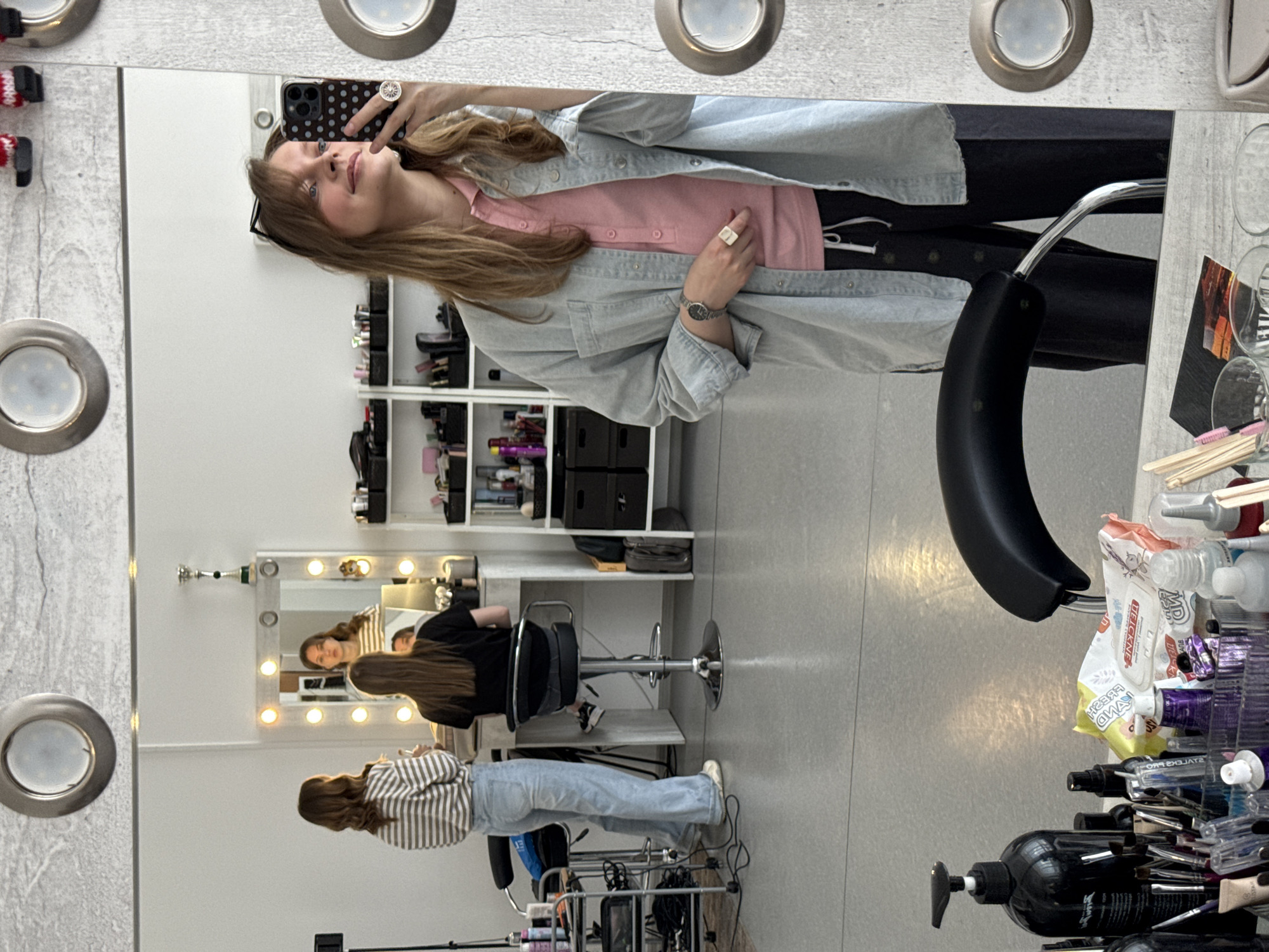 A woman is taking a selfie in a mirror in what appears to be a hair salon. She is wearing a pink shirt, a light blue denim jacket, and black pants. The mirror has lights around the edges. In the reflection, there are two other people: one is sitting in a salon chair getting their hair done, and the other is standing and appears to be working at the salon. There are shelves with various hair products in the background. There are hair styling tools and products on a table in the foreground. The image does not provide information about the city of Izhevsk, Russia.