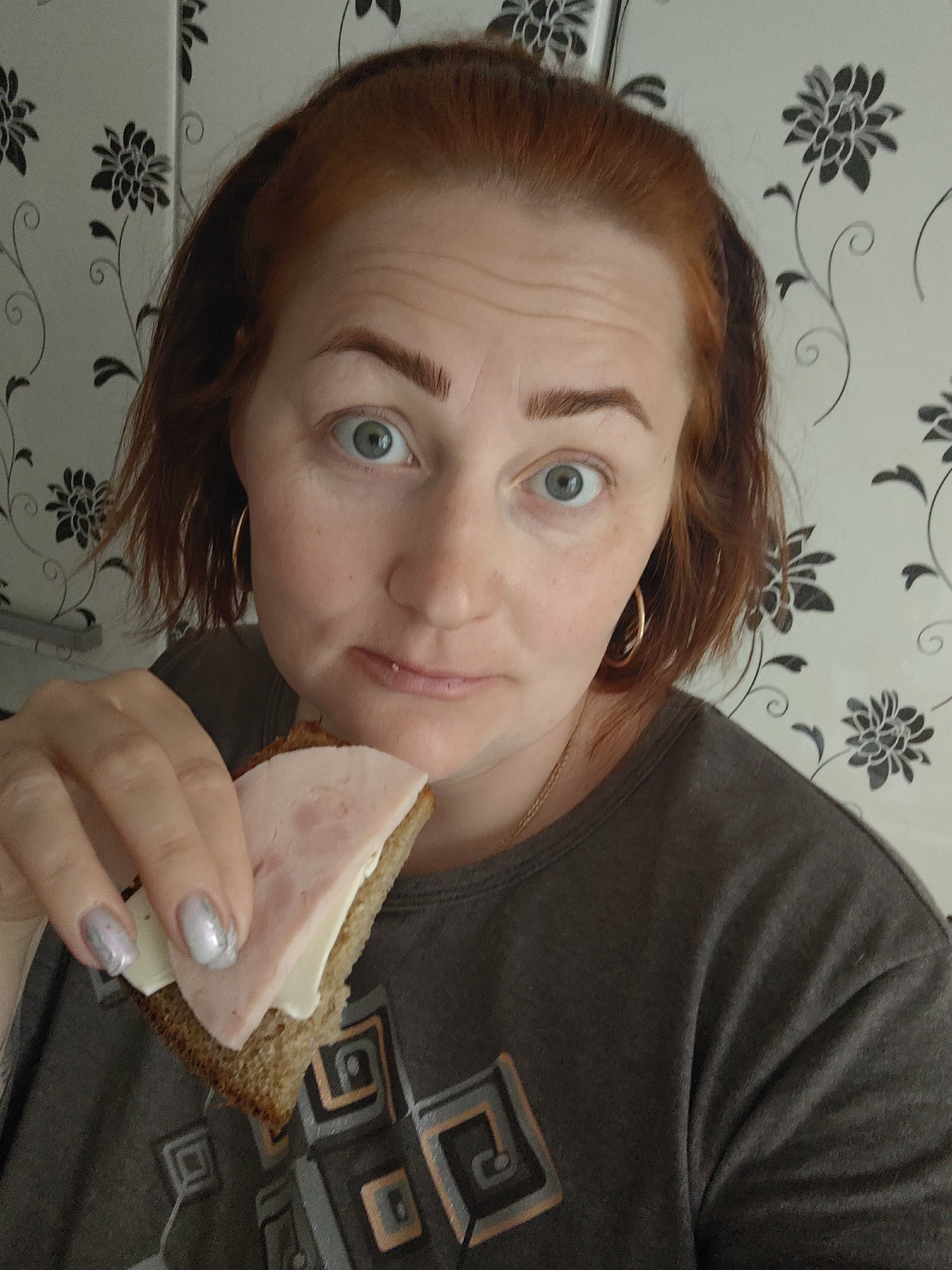 A woman with red hair is holding a sandwich with ham and cheese. She is wearing a gray t-shirt and gold hoop earrings. The background is a white wall with a black floral pattern. The location is not discernible as Фоки, Russia.