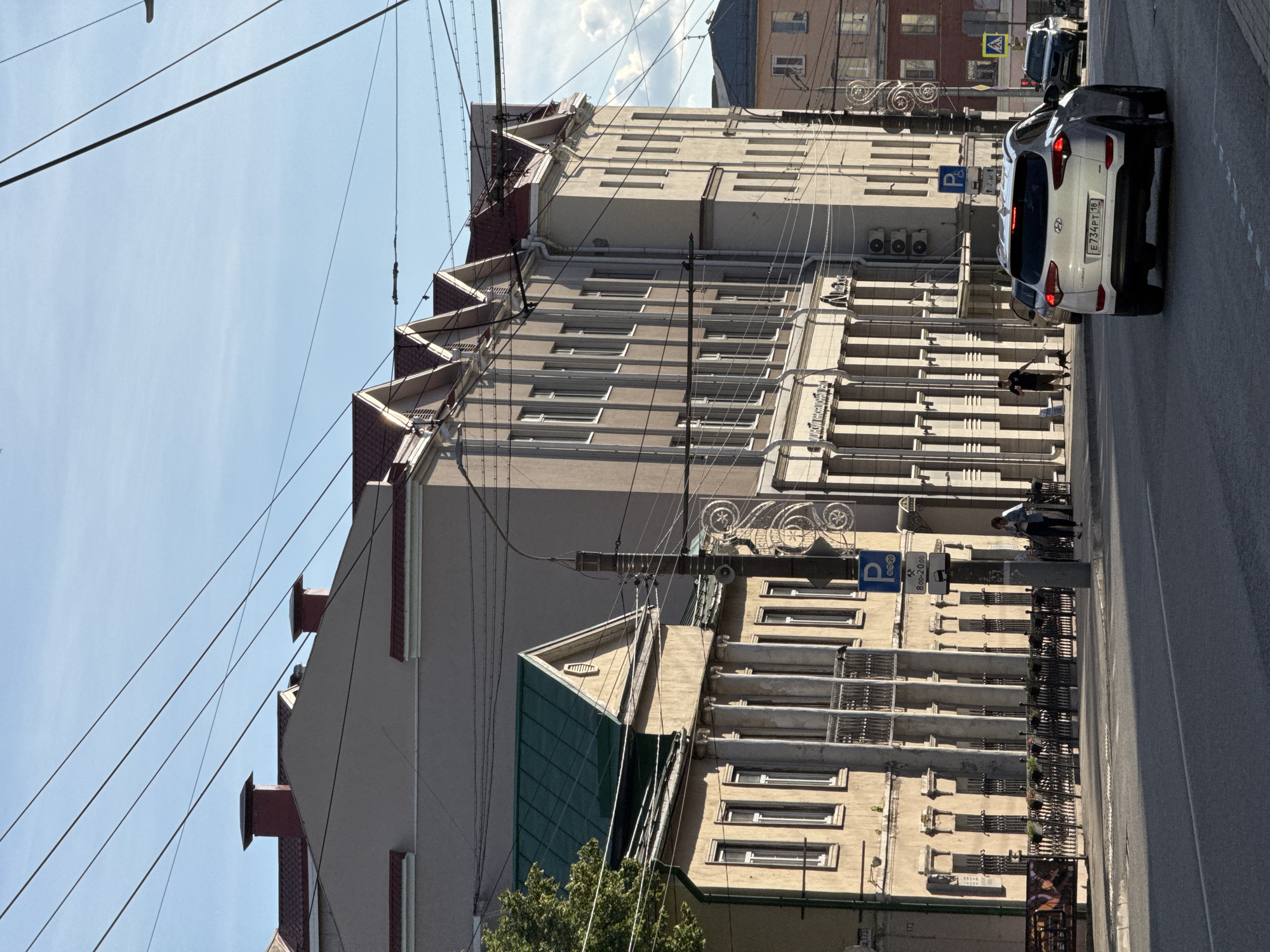 The image depicts a street scene in Izhevsk, Russia, likely during daylight hours. In the foreground, a white Hyundai SUV is driving on a paved road. The background features several buildings with distinct architectural styles. The building on the left has a light-colored facade, a green sloped roof, and classical architectural elements like columns. Adjacent to it, a larger building dominates the scene with a lighter facade, multiple stories, and a stepped roofline with red tiles. Overhead, a complex network of utility wires crisscrosses the sky. A street lamp with decorative elements is visible near the sidewalk. A few individuals are present on the sidewalk, including a person walking a dog. Traffic signs, including a parking sign and a time-restricted parking sign, are also visible. The sky is partly cloudy, with a clear blue expanse visible in the upper portion of the image.
