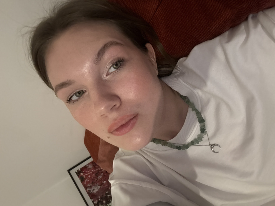 A young adult female is depicted in a close-up, tilted, indoor photograph, looking directly at the camera. She has fair skin, light brown hair, and green eyes, with a small mole positioned superior to the right side of her upper lip. She wears a white long-sleeved top. Her accessories include a necklace constructed from green, irregularly shaped stone beads, and a second, longer silver-colored necklace featuring a crescent moon pendant. The subject is partially reclined against a reddish-orange textured surface, consistent with a sofa or cushion. In the background, a dark-framed artwork with abstract red and white patterns is visible on a light-colored wall. The scene is illuminated by indoor artificial lighting. This location is within the Vozdovac Urban Municipality, Serbia.