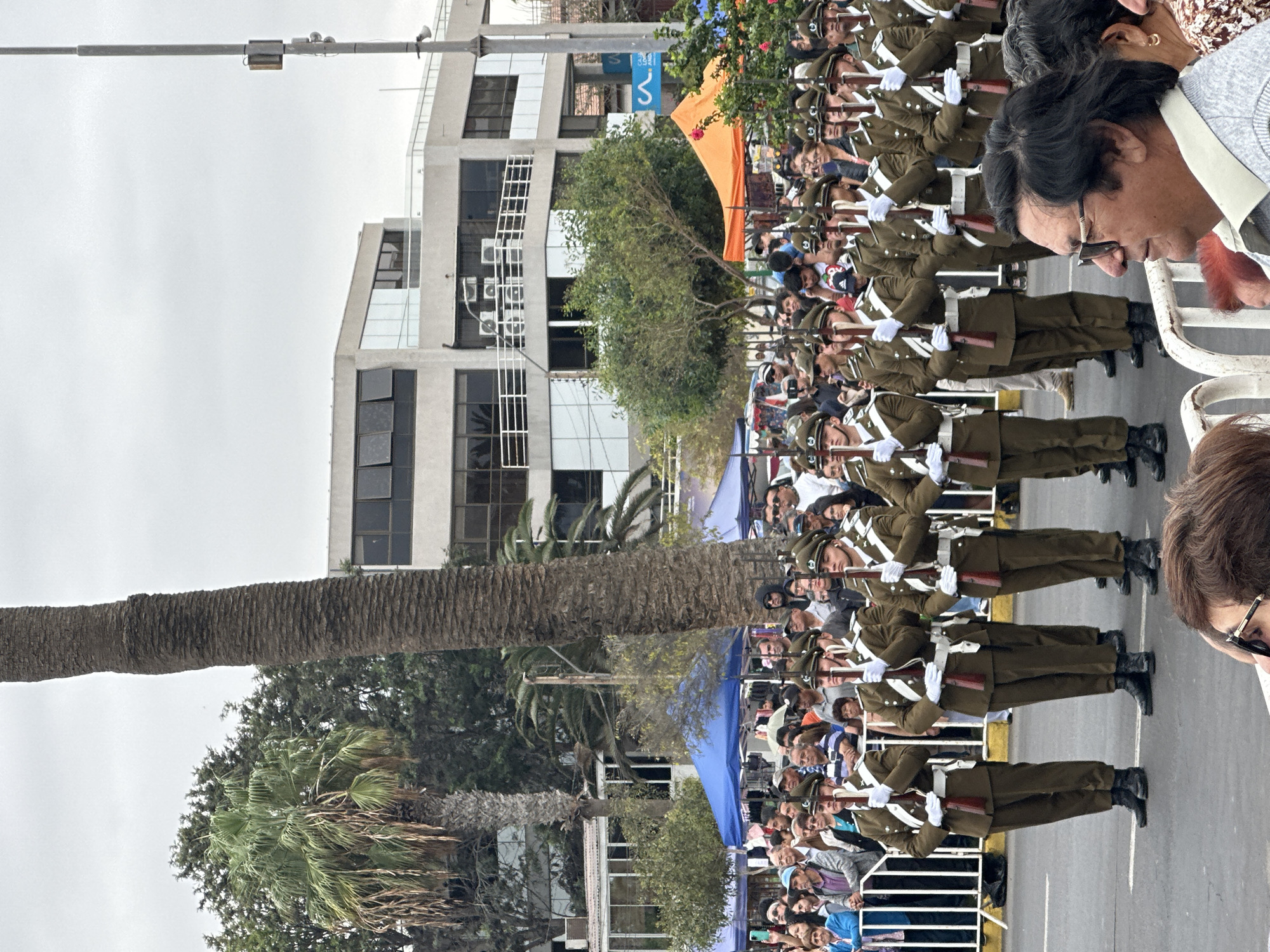 A scene in Arica, Chile, depicts a military parade. Soldiers in olive-green uniforms and white gloves march in formation, each holding a rifle. Behind them, spectators line the street, with tents and barriers visible. The backdrop includes urban architecture, palm trees, and an overcast sky.