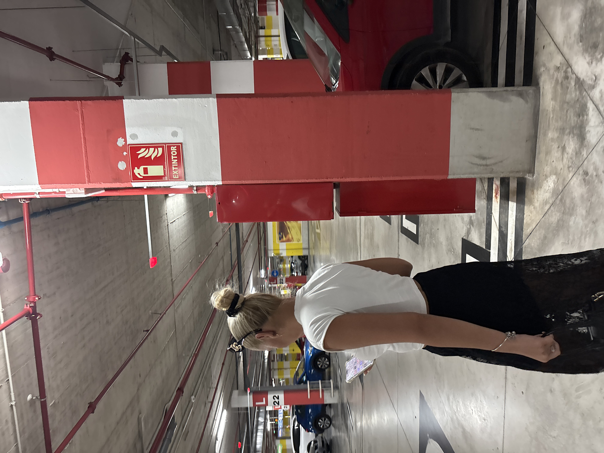The image shows a blonde woman in a white crop top and black lace skirt holding a phone in a parking garage in Orihuela, Spain. She is standing in front of a red and white striped pillar with a fire extinguisher sign. Several cars are visible in the background, including a red one on the right. The environment appears to be an underground parking structure with concrete walls and ceiling, illuminated by overhead lighting.