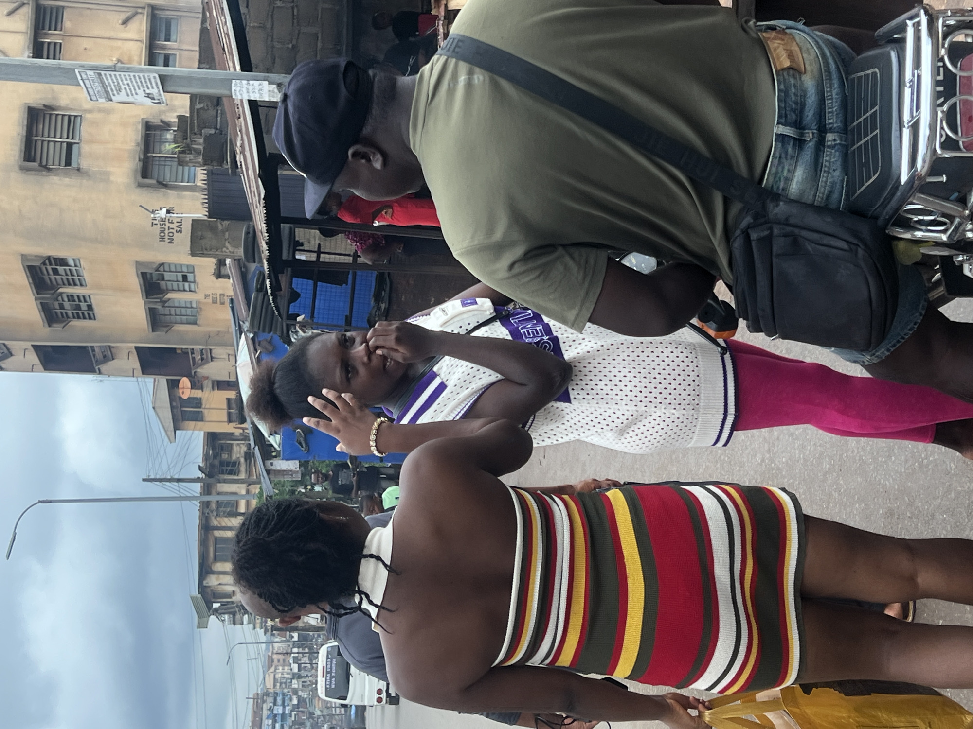 The image depicts a street scene in Nkpor, Nigeria, with several individuals and buildings visible. In the foreground, three people are the primary subjects. On the left, a woman faces away from the camera, wearing a strapless, horizontally striped dress in red, yellow, green, and white. She carries a yellow shopping bag. To her right, a young woman with her hair in a bun is seen from the chest up, facing the camera and covering her mouth with her hand. She wears a white sleeveless top with a pattern of small dots and a white collar. A gold bracelet adorns her wrist. To the right, a man wearing a black baseball cap and an olive green t-shirt faces away from the camera, looking towards the young woman. He has a black crossbody bag slung over his shoulder and is seated, possibly on a motorcycle, as indicated by a portion of a vehicle visible beneath him, which includes chrome accents and a textured seat.
In the background, the environment suggests a bustling urban setting. Buildings, some appearing aged and weathered with visible signs of wear and tear, line the street. One building has a sign that reads "THIS HOUSE IS NOT FOR SALE." A white van is parked on the left side of the street, and in the distance, other vehicles and more buildings are visible, contributing to the impression of a busy road. The sky is overcast, suggesting a cloudy day. The overall atmosphere is that of a typical day in a Nigerian city, with people going about their activities amidst the urban landscape.