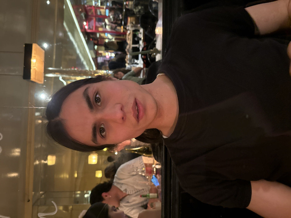 A person with long brown hair and brown eyes wearing a black shirt faces the camera. A mirror behind reflects restaurant activity and lighting. A fair-skinned woman in a white shirt sits at a table in the lower portion of the image, appearing to be in conversation. The scene suggests a dining environment.