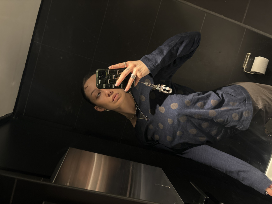 A female subject is depicted in a mirror selfie taken within a dark-tiled indoor environment, likely a restroom. She holds a dark green smartphone, featuring a textured case and an attached beaded charm, in her left hand to capture her reflection. The subject wears a long-sleeved navy blue top with large, faded grey polka dots, dark-colored trousers, small hoop earrings, a silver chain necklace with a small pendant, and rings on her fingers. Her hair is pulled back. The background shows large, dark, reflective wall tiles, with a metallic fixture (possibly a dispenser) on the left and a toilet paper holder with a white roll on the lower right wall. The specific location within London, United Kingdom, is not discernible from the visual evidence.