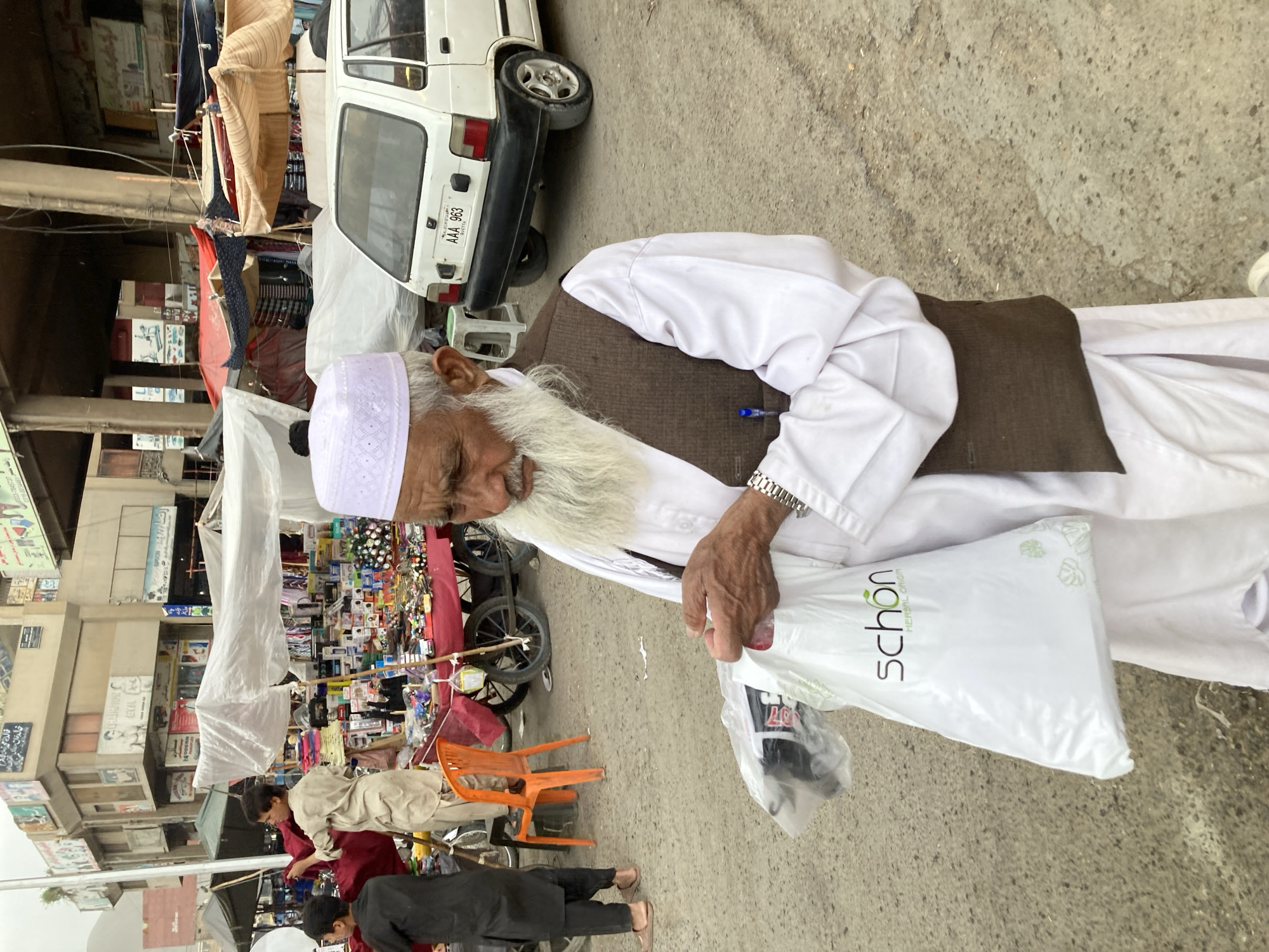 An older man with a long white beard, wearing a white top, brown vest, and white skullcap, stands on a street in Quetta City Tehsil, Pakistan, holding a white shopping bag with the word "schön" printed on it. The man is also wearing a silver watch. Behind him, a street market is visible with various items on display. People are present in the market area. A white car is parked to the man's left. The street is paved. The general environment suggests a bustling urban market scene.