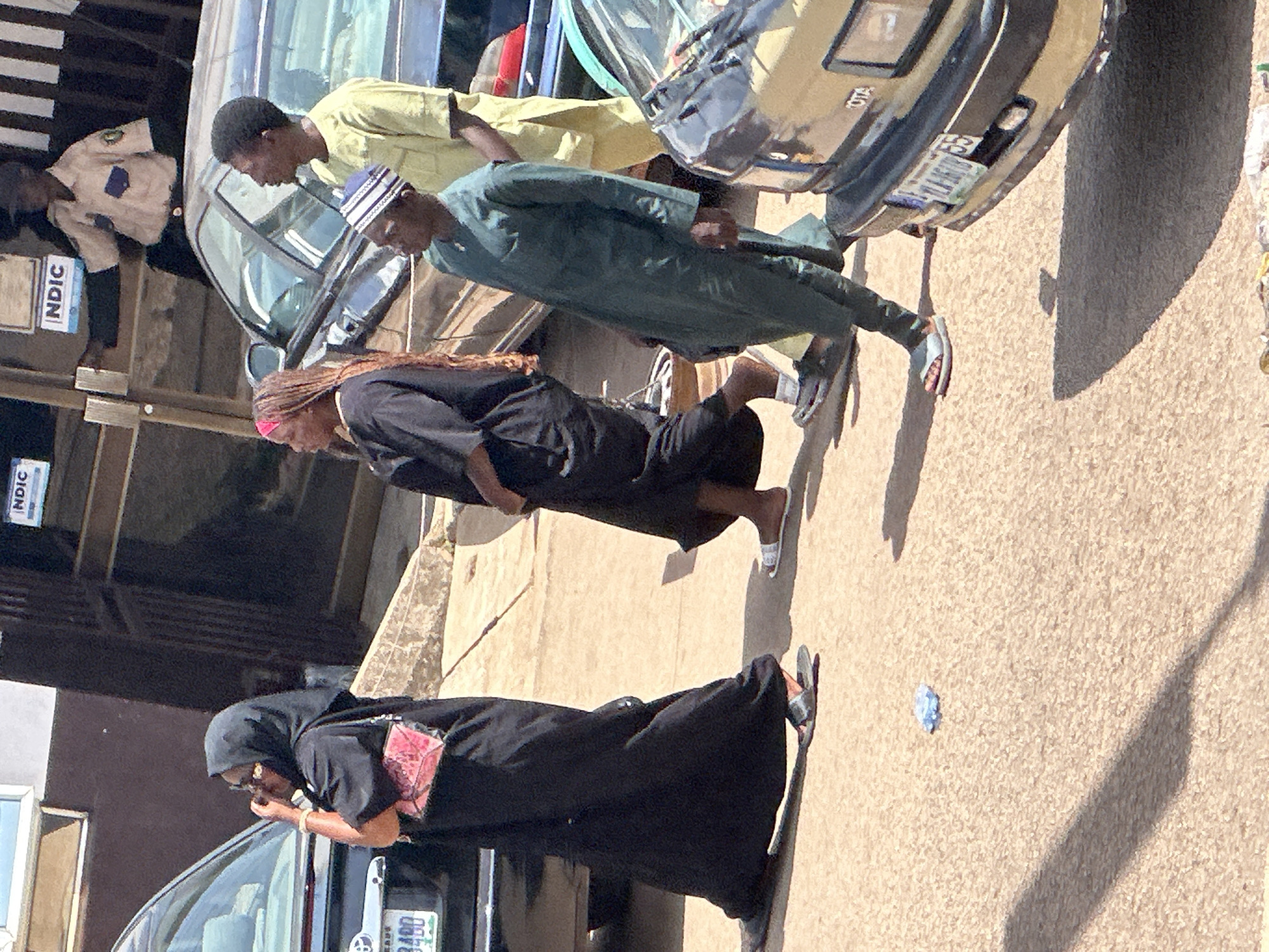 In Bukuru, Nigeria, on a sunny day, several individuals are captured in a street scene. To the left, a person wearing a black hijab and abaya, with a pink patterned handbag slung over their shoulder, walks to the left of the frame, wearing sandals. Adjacent to them, a woman with a pink headband and long braids, dressed in black, walks towards the right, also in sandals. In the center, a man in a light yellow garment and dark trousers walks to the right, while a man in a dark green traditional outfit and a striped cap walks in the foreground to the right, wearing sandals. Behind them, a security guard in a tan uniform is visible near a building entrance. Various vehicles, including a dark sedan on the left and a light-colored Toyota sedan on the right, are present. The ground is a textured, sandy surface, with a small blue object lying on it in the foreground. Shadows are cast on the ground, indicating the sun's position. The environment suggests an urban setting, likely a street or parking area.