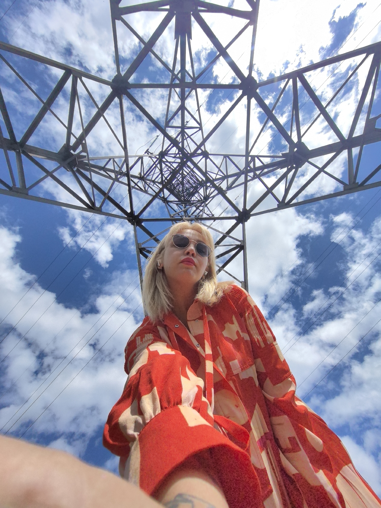 An extreme low-angle, wide shot captures a blonde woman with sunglasses and bright pink lipstick looking up from below a towering metal pylon in Noviy, Russia. The woman is positioned in the lower right of the frame, her upper body visible from the chest up, wearing a loose-fitting orange and white patterned shirt with voluminous sleeves. Her eyes are hidden behind round, dark sunglasses. The pylon, a complex lattice of metal beams, dominates the upper half of the image, its structure receding into the distance and framing the bright blue sky dotted with white clouds. Several high-voltage power lines are visible crisscrossing the sky behind the pylon. The perspective suggests the photograph was taken looking directly upwards from the base of the pylon.