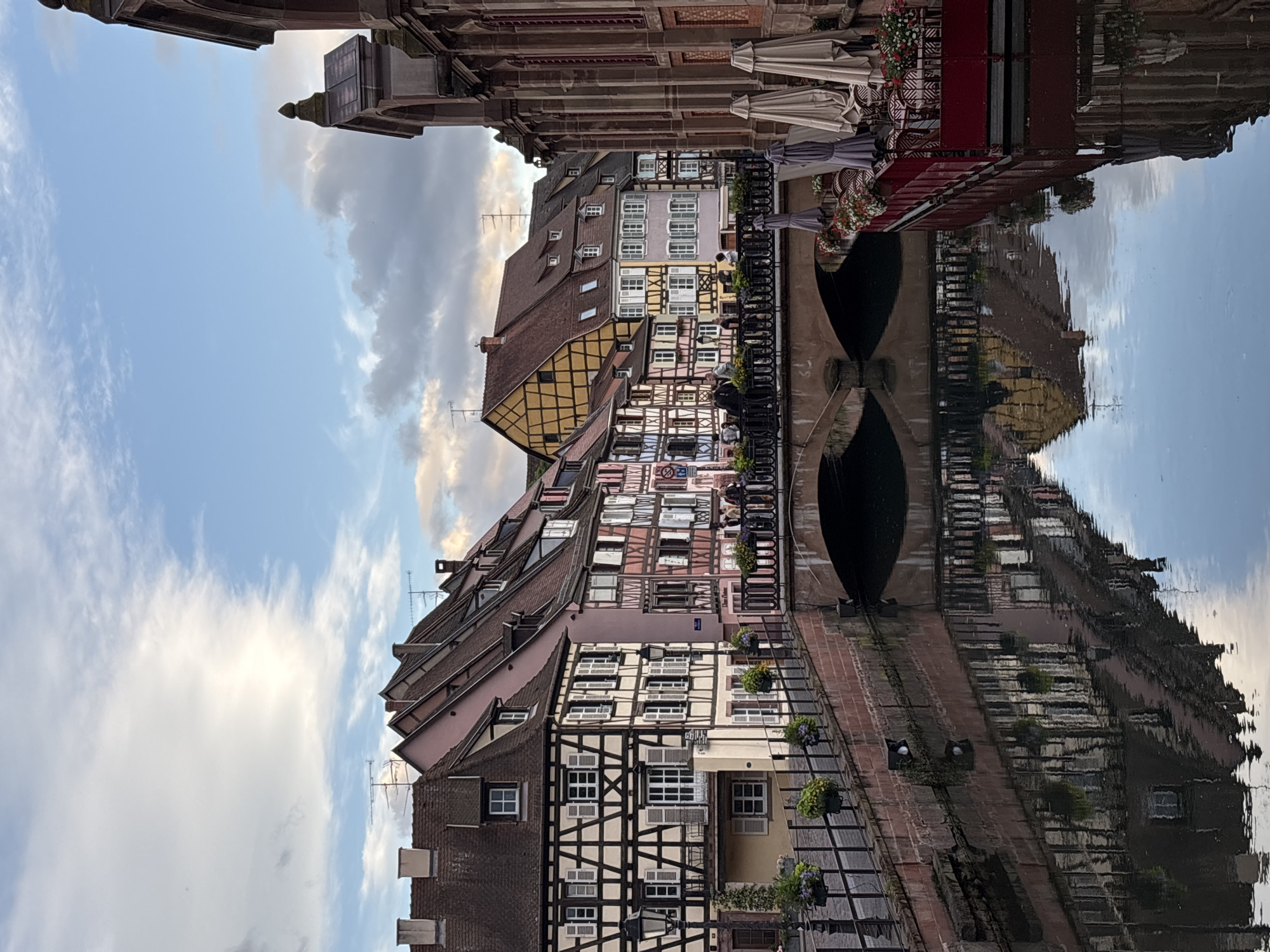 The image depicts a picturesque canal in the Petite Venise district of Colmar, France. The canal is flanked by colorful half-timbered buildings with steeply pitched roofs. The architecture suggests a German influence. A stone bridge crosses the canal, and people are visible walking across it. On the right side of the canal, a red-colored structure with umbrellas and tables suggests an outdoor seating area for a restaurant or cafe. The water reflects the buildings and the sky, creating a mirror-like effect. The sky above is partly cloudy with streaks of white clouds against a blue background. The overall scene evokes a charming and tranquil atmosphere typical of European canal towns.