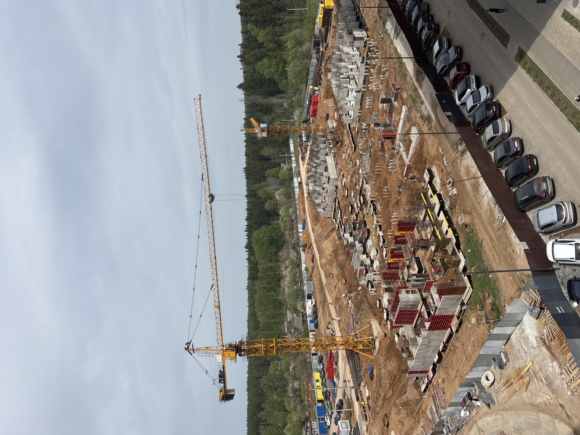 The image shows a construction site in Izhevsk, Russia. A large yellow construction crane dominates the view. The site contains the foundations of buildings, with wooden and metal forms outlining the shapes of walls and rooms. Stacks of concrete blocks and other building materials are present. The surrounding area includes a forest and a parking lot filled with cars. No people are clearly visible, though there may be individuals present on the construction site.